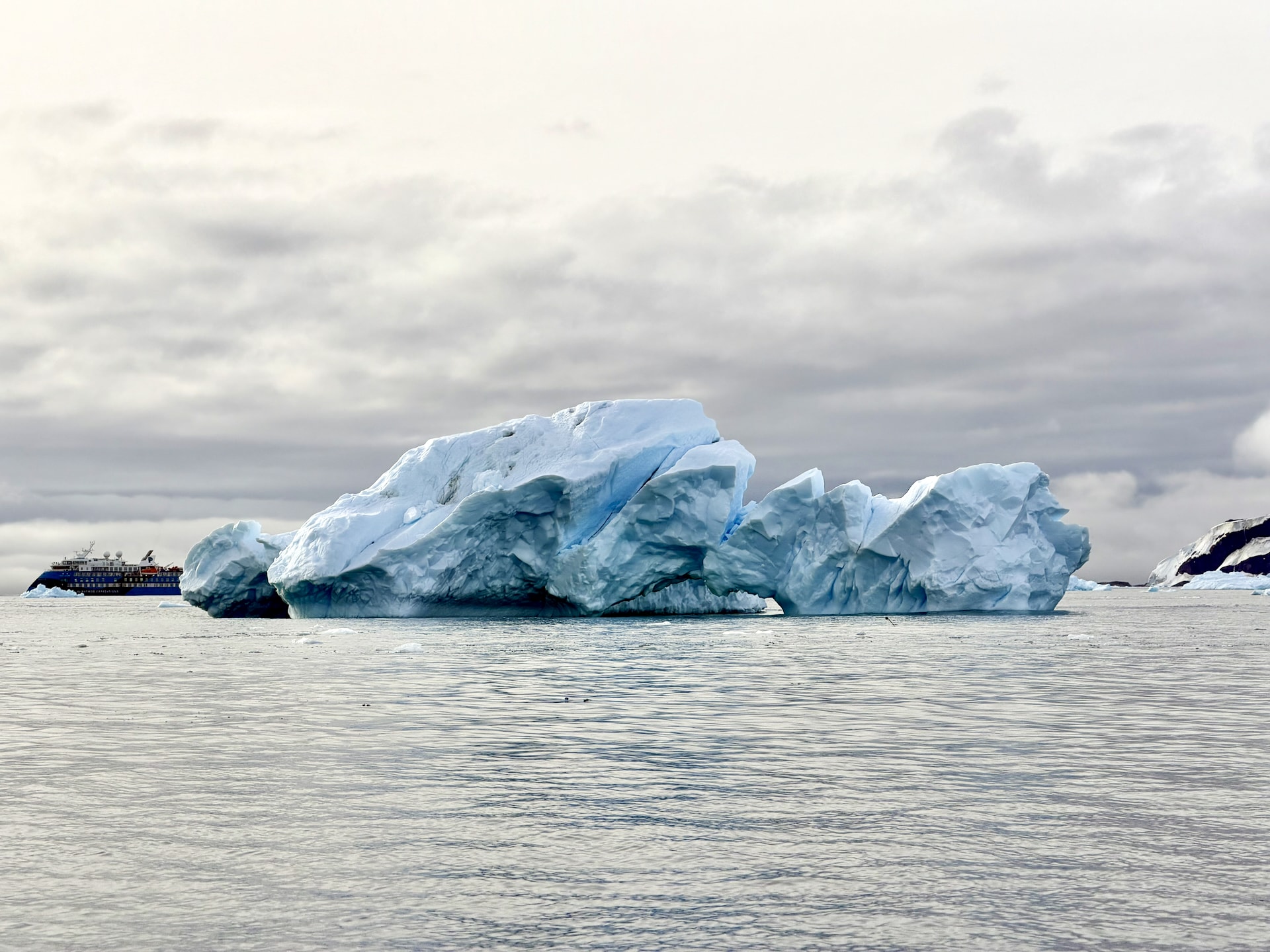 A large blue-white iceberg in the foreground with the Ocean Albatros expedition ship visible in the distance behind it, under an overcast Antarctic sky.