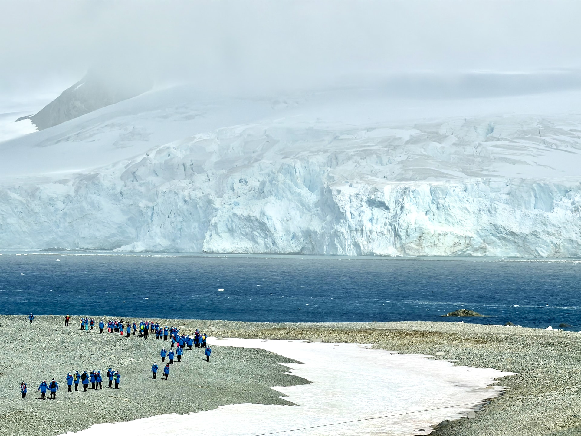 Expedition members walking on Half Moon Island Antarctica Group of expedition passengers in blue parkas walking on a gravel beach at Half Moon Island, with glaciers and mountains across the water.