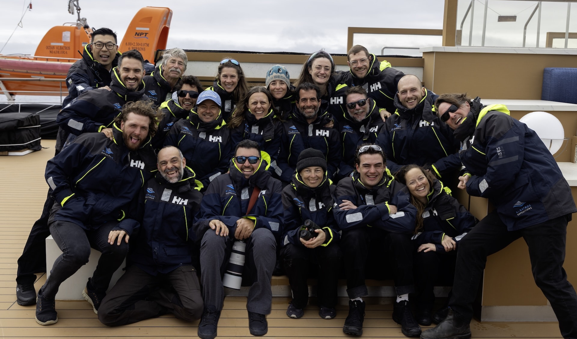Ocean Albatros expedition team group photo Group photo of approximately twenty expedition team members in matching navy Helly Hansen jackets, posing together on the deck of the Ocean Albatros with orange lifeboats visible behind them.