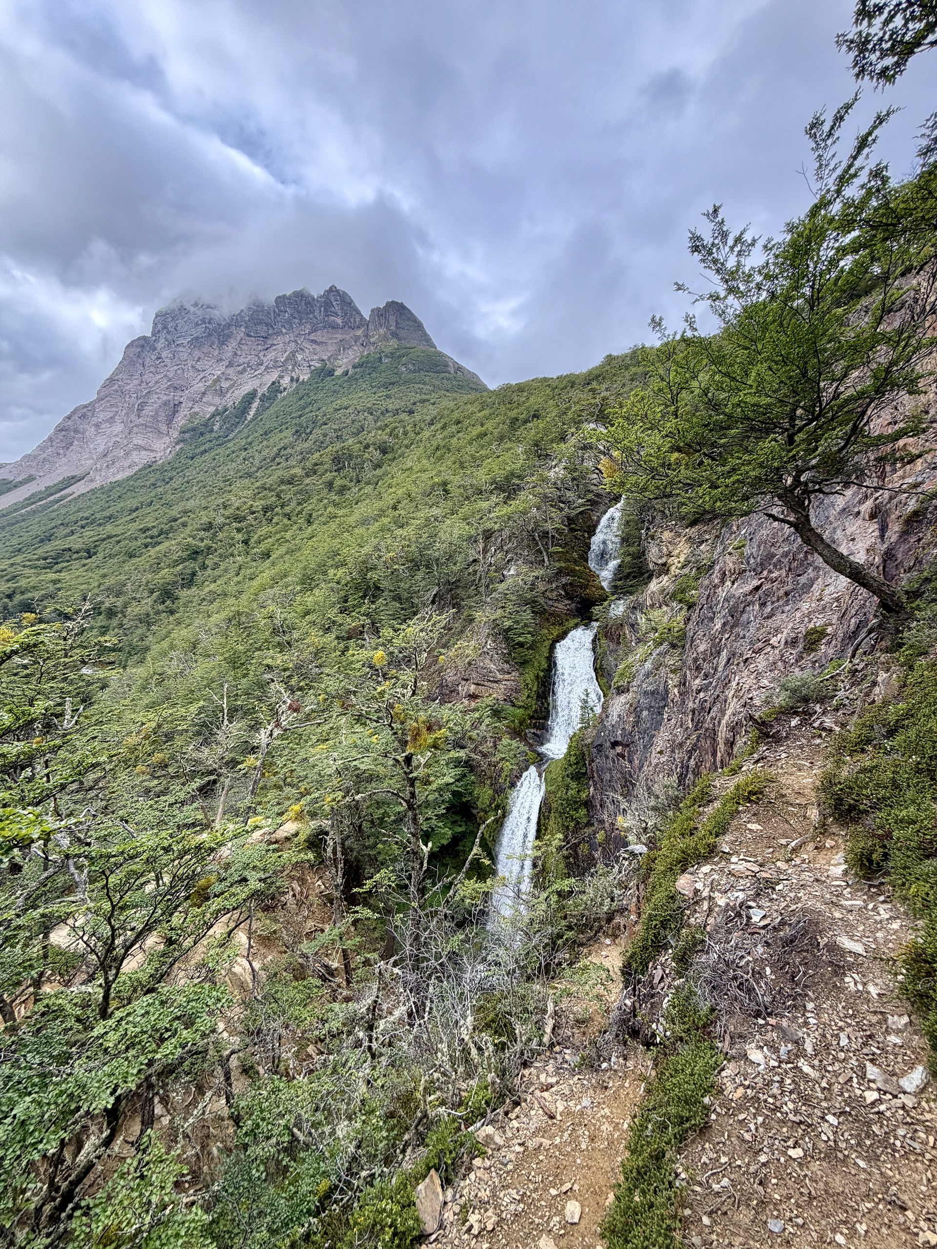 Waterfall and jagged peaks on hiking trail Patagonia Argentina Waterfall cascading down rocky mountainside with jagged Patagonian peaks and hiking trail in the foreground