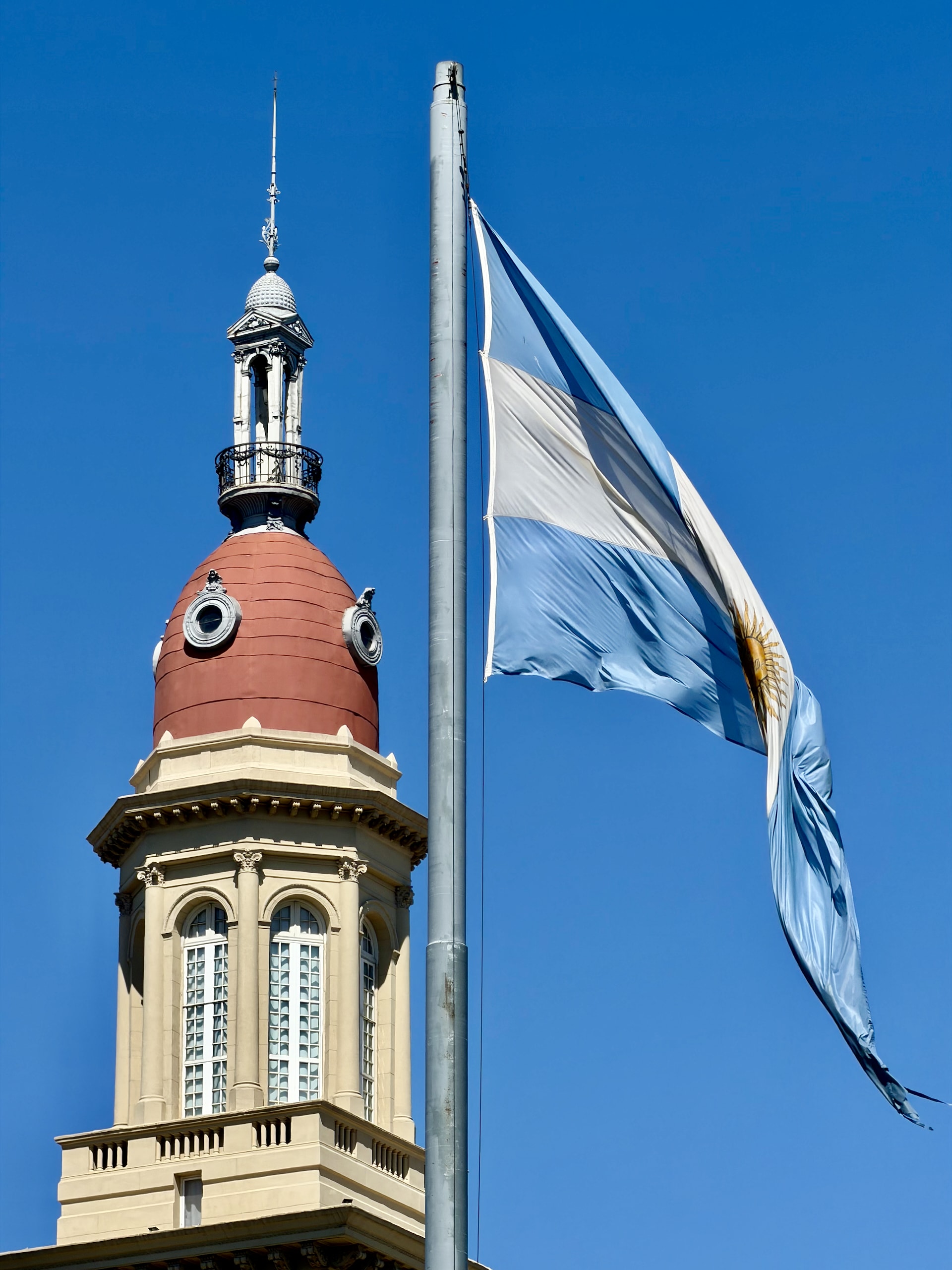 The Argentine flag flying beside the domed tower of a historic Buenos Aires building against a clear blue sky.