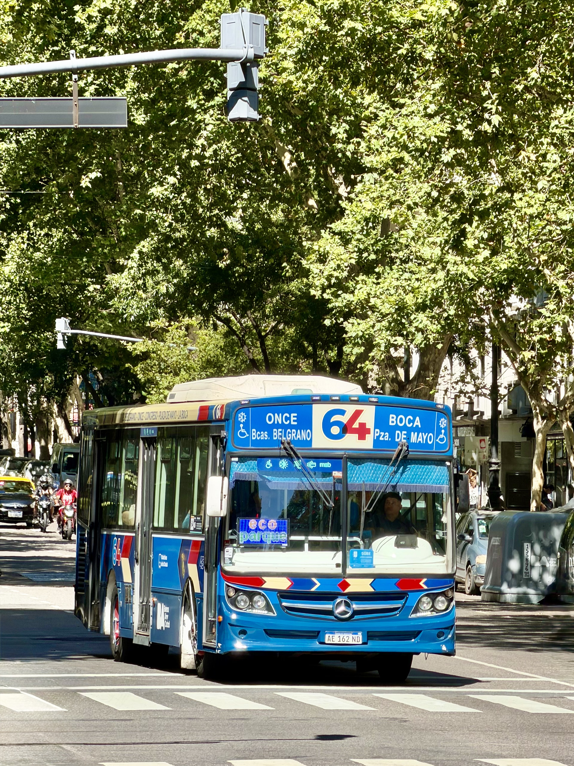A blue colectivo bus on line 64 (Once–La Boca) driving down a tree-lined Buenos Aires avenue.