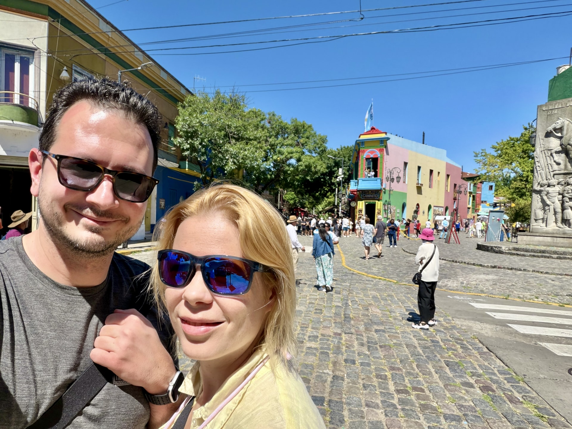 Laszlo and Andrea taking a selfie on the cobbled Caminito street in La Boca, colorful buildings behind them.