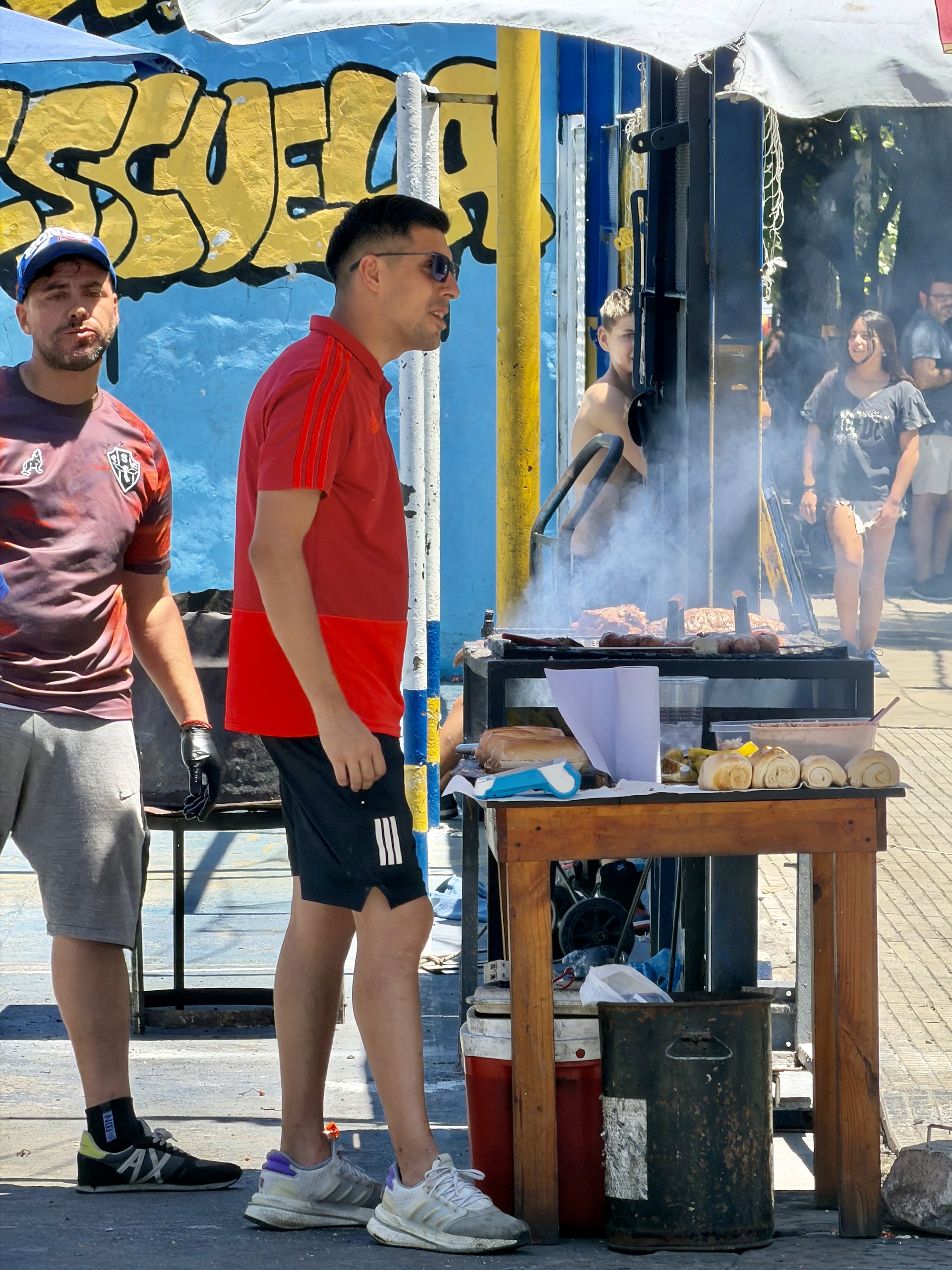 Choripan being grilled near La Bombonera stadium Sausages smoking on a grill with bread rolls waiting nearby, a choripan stand near La Bombonera stadium.