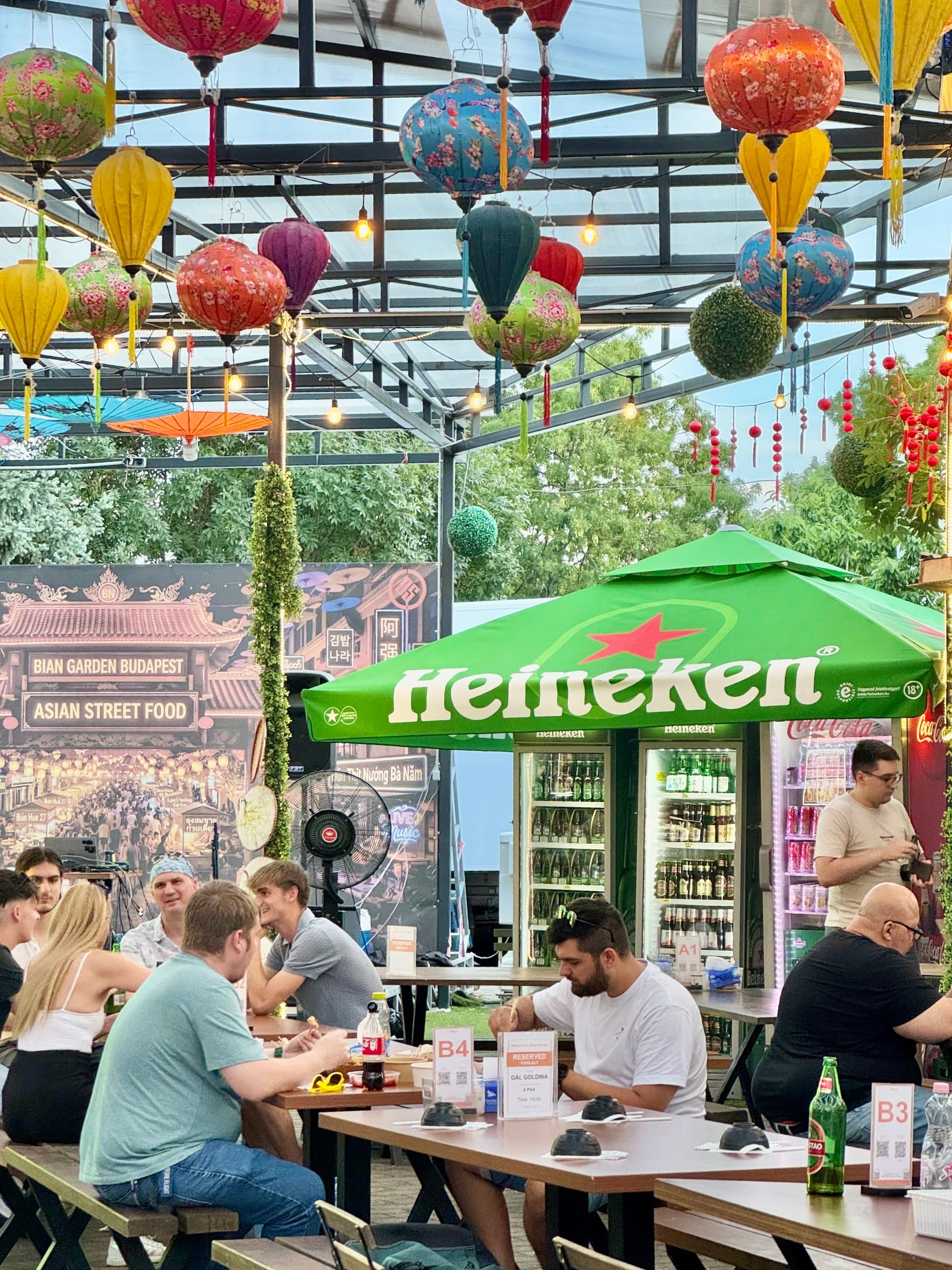 Covered seating area at Bian Garden Budapest Guests at long tables under the high lantern-hung roof at Bian Garden, with a drinks fridge behind