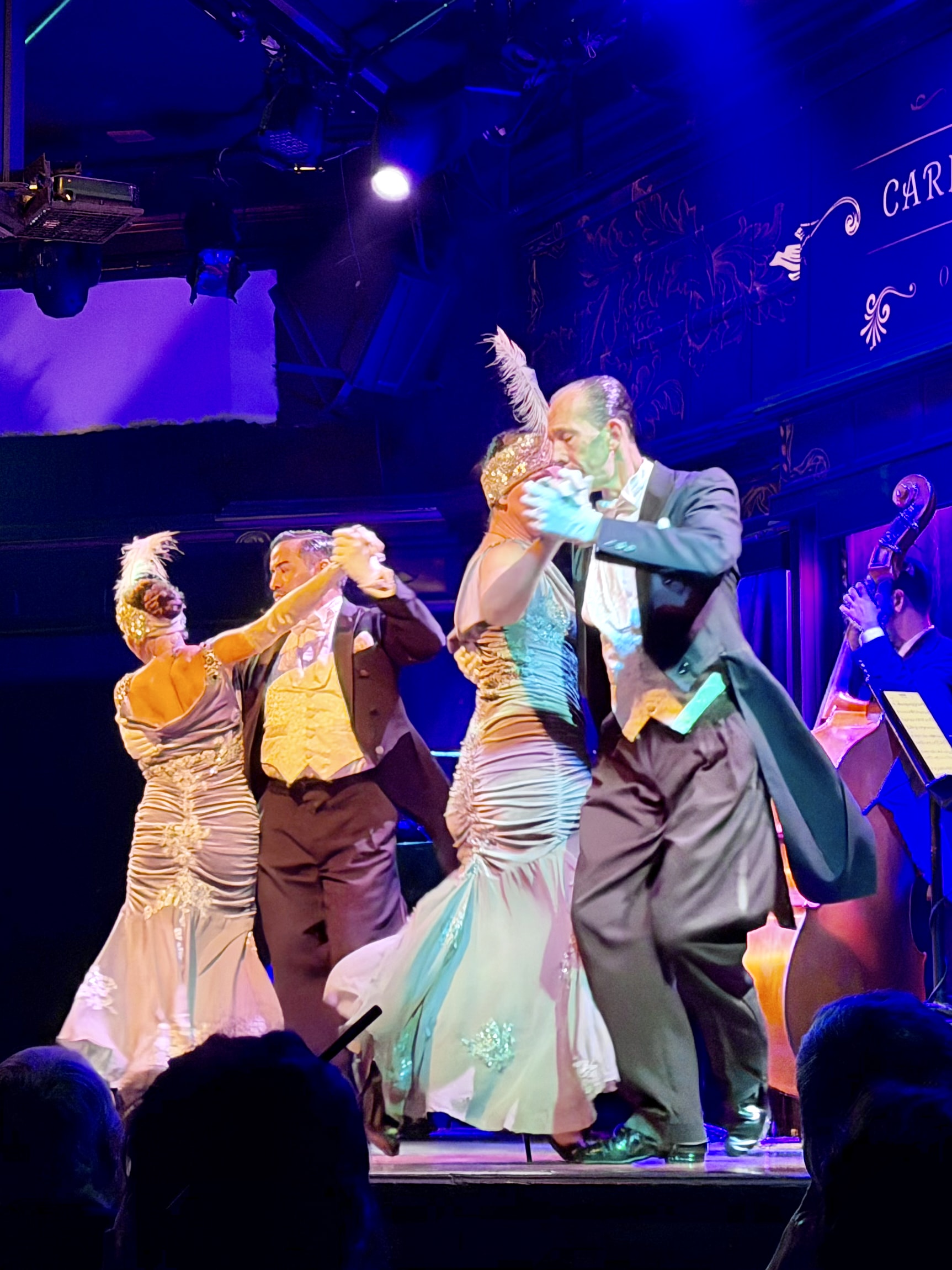 Dancing to Carlos Gardel at the El Querandí tango show Two couples in elegant 1930s formalwear dancing tango at El Querandí, in front of a Carlos Gardel-themed backdrop.