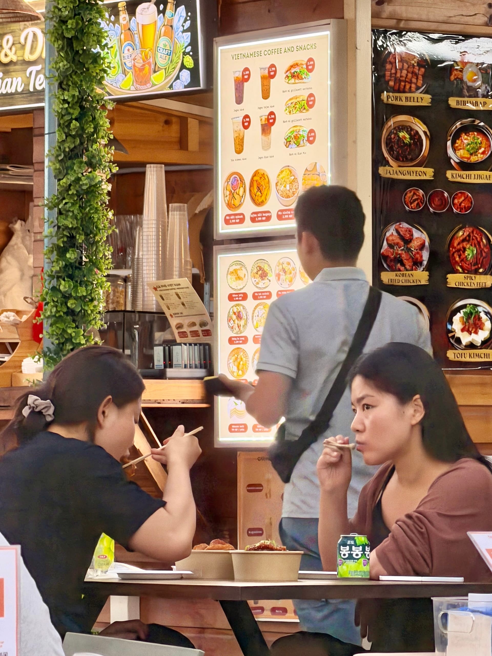 Guests eating at Bian Garden Budapest Two women eating with chopsticks in front of Vietnamese coffee and Korean menu boards at Bian Garden
