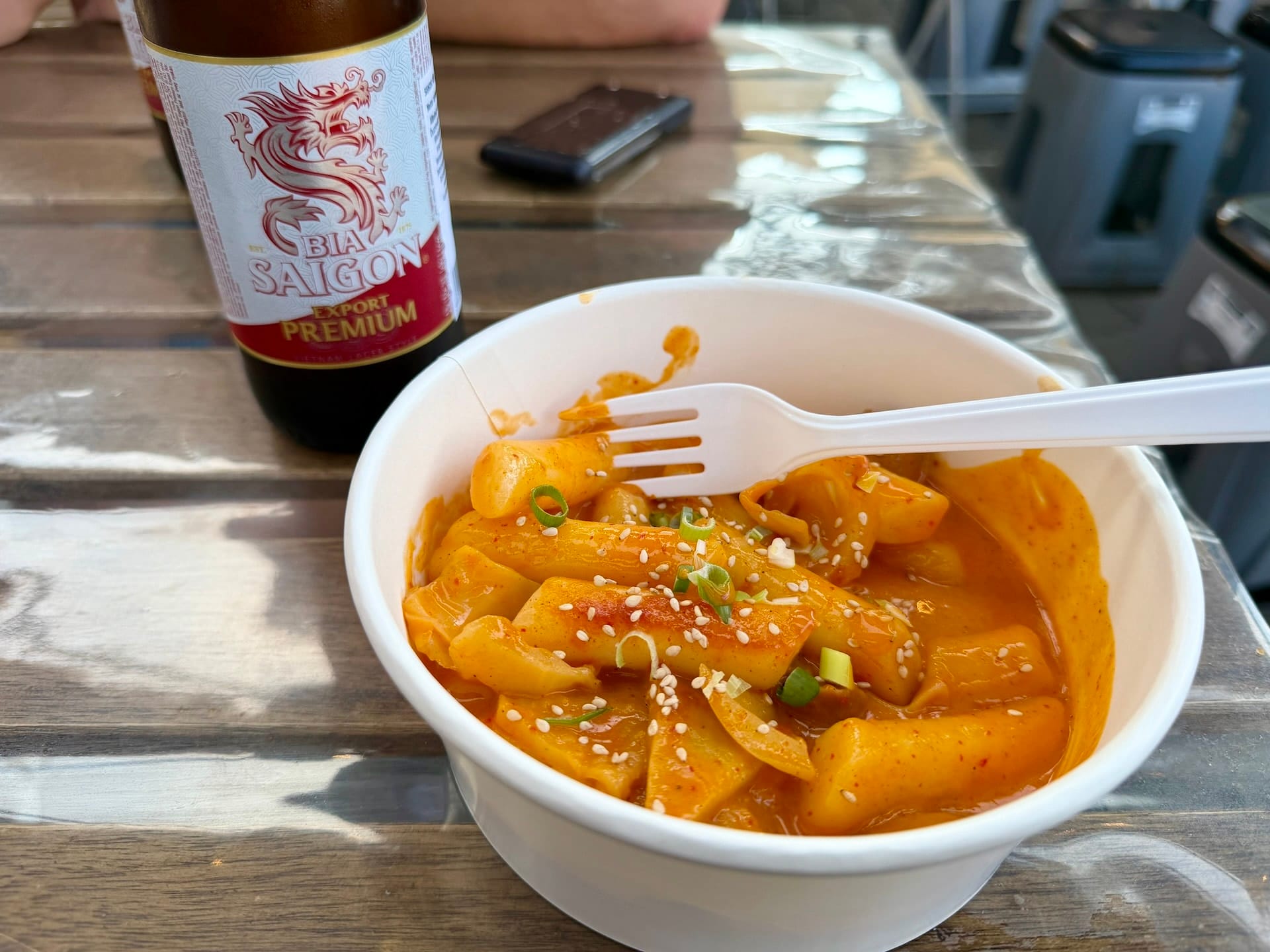 Korean tteokbokki with Bia Saigon beer at Bian Garden Bowl of tteokbokki in red sauce with sesame seeds and scallions, next to a bottle of Bia Saigon beer