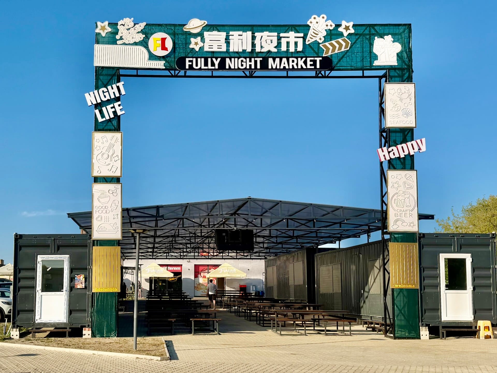 Fully Night Market entrance and closed containers in daylight The Fully Night Market entrance gate in daylight, with closed shipping-container stalls and empty benches behind it.