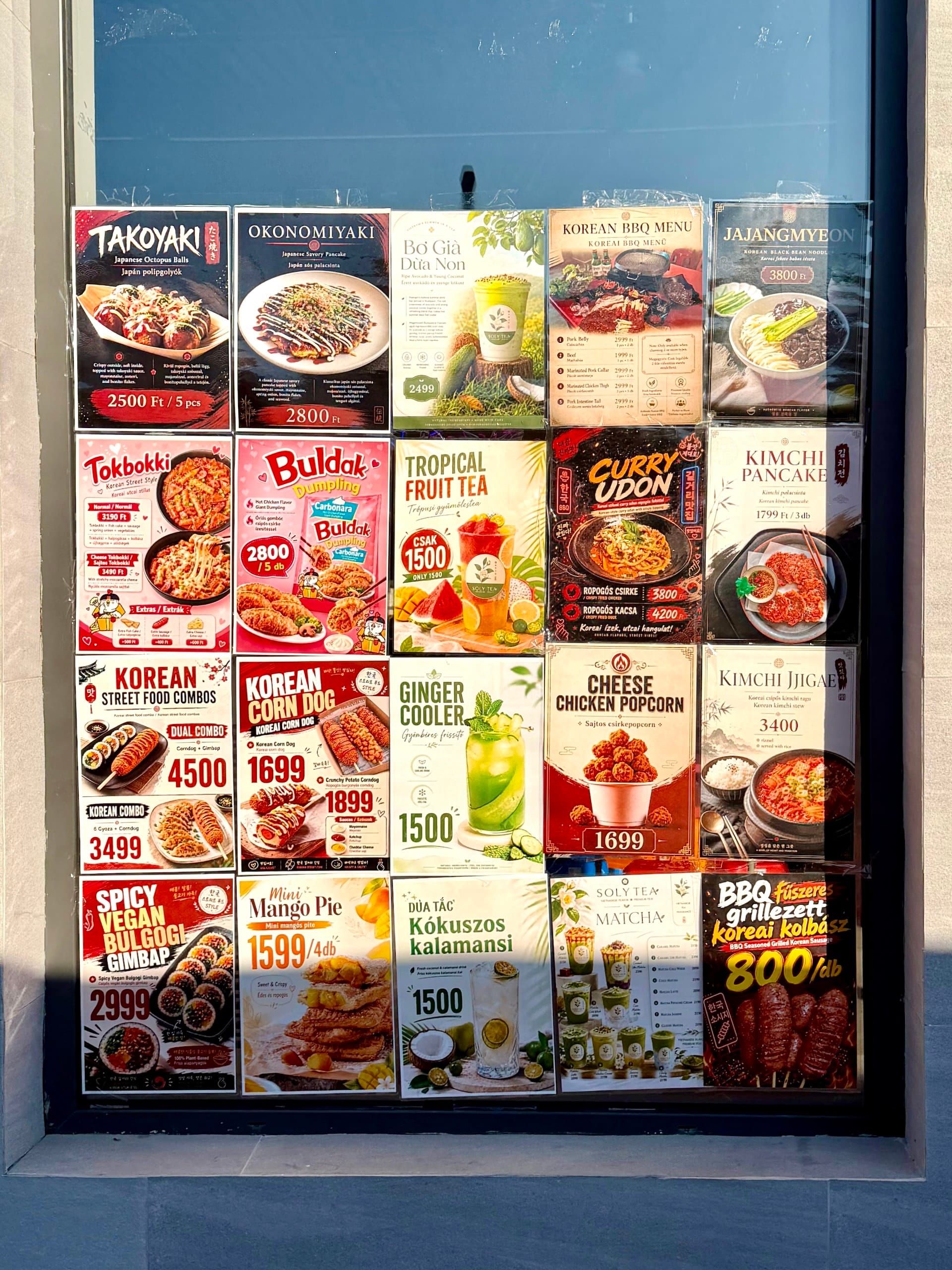 Japanese and Korean menu posters at Fully Night Market Budapest Menu posters for takoyaki, okonomiyaki, Korean BBQ, tteokbokki and corn dogs in a stall window at Fully Night Market.