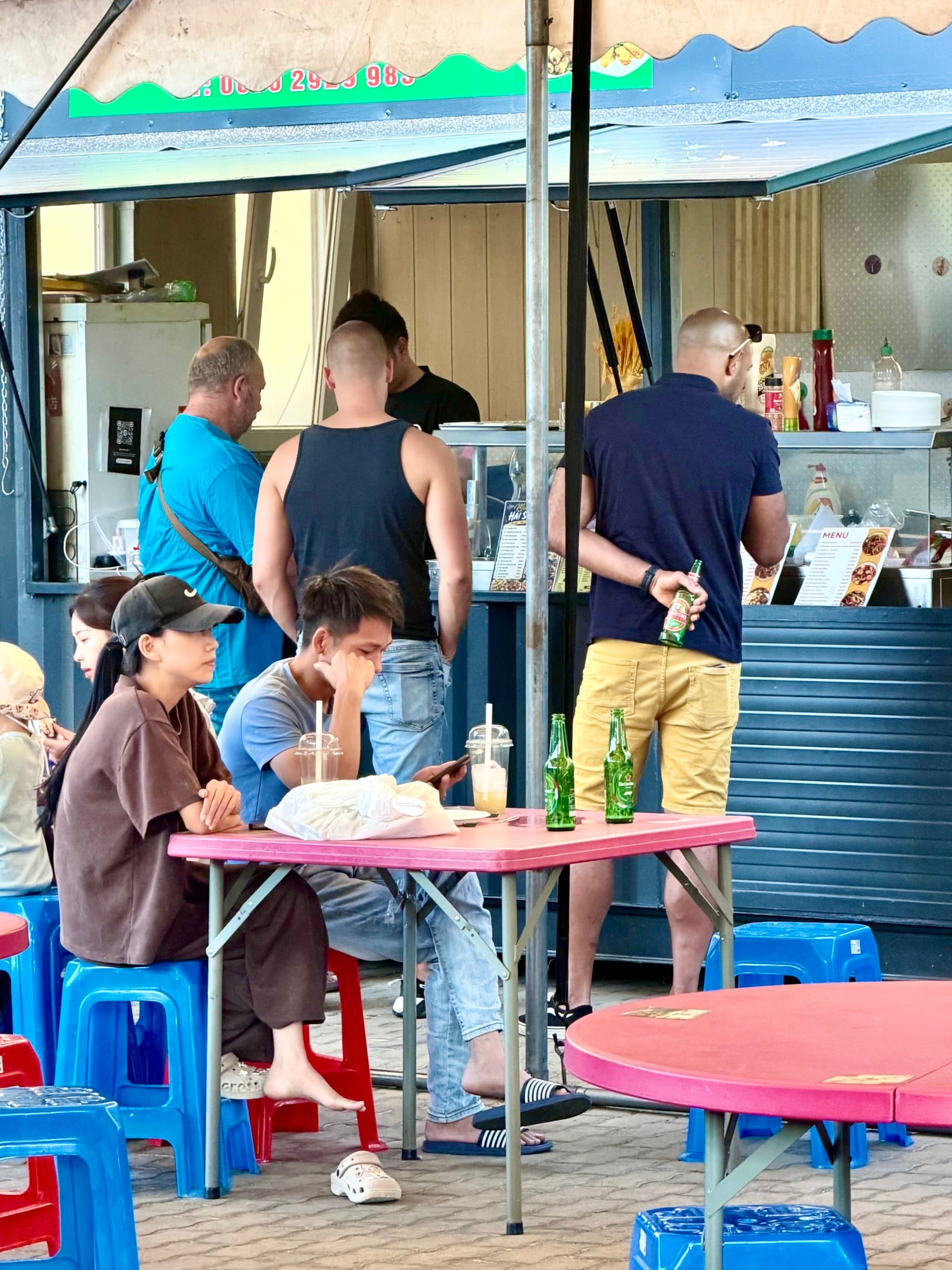 Plastic stools and tables at Fully Night Market Budapest Guests on low blue plastic stools at pink tables at Fully Night Market, with a food stall behind them.