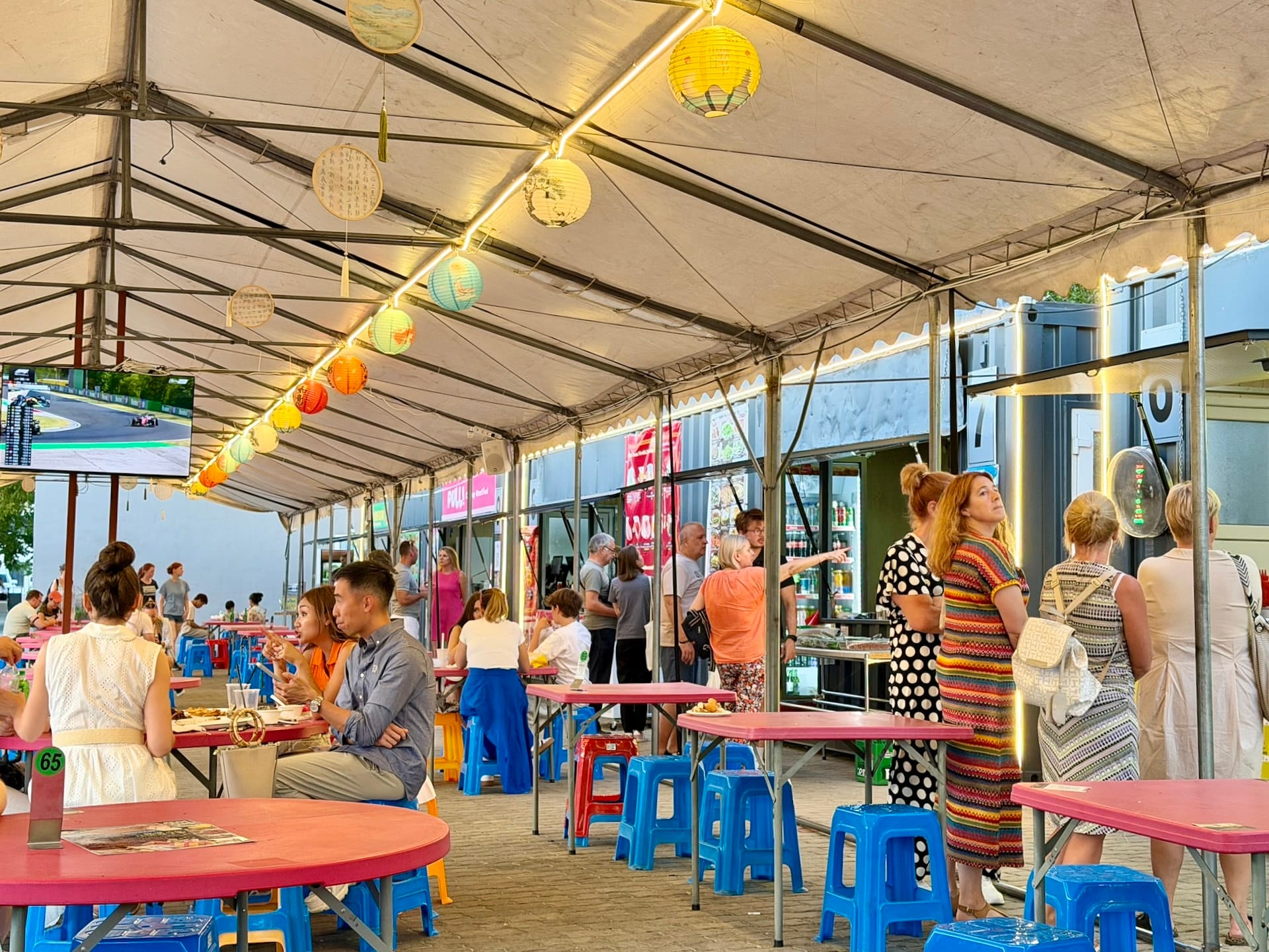 The largest open-sided tent at Fully Night Market Budapest The largest tent at Fully Night Market, open along one side, with paper lanterns overhead and a race playing on the screen.