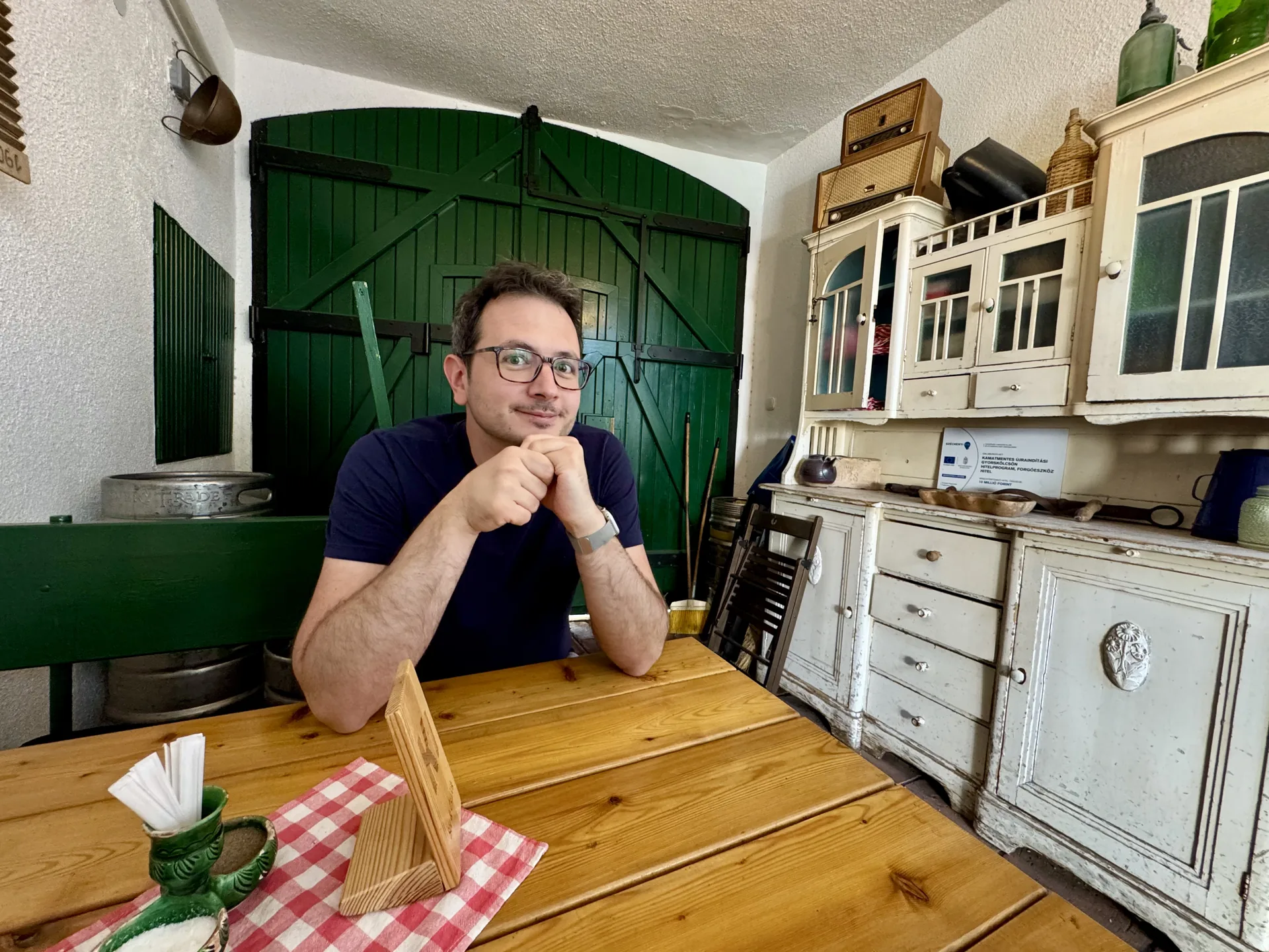 Lunch at the Zöldkapu after the caves, Budapest Laszlo sitting at a wooden table at the Zöldkapu, green barn doors and a painted dresser behind him