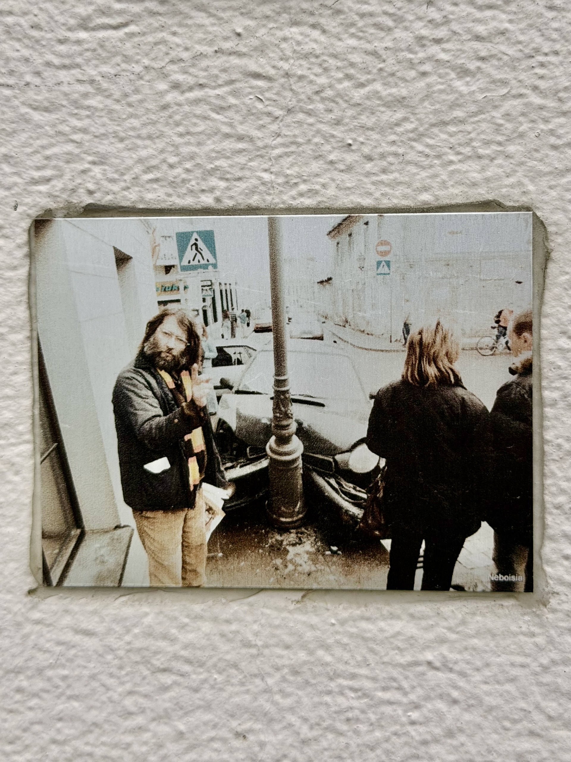 Archival photo of a car crash in Užupis An archival photograph mounted on a white wall in Užupis showing a car that has hit a lamp post, a bearded man standing beside it.