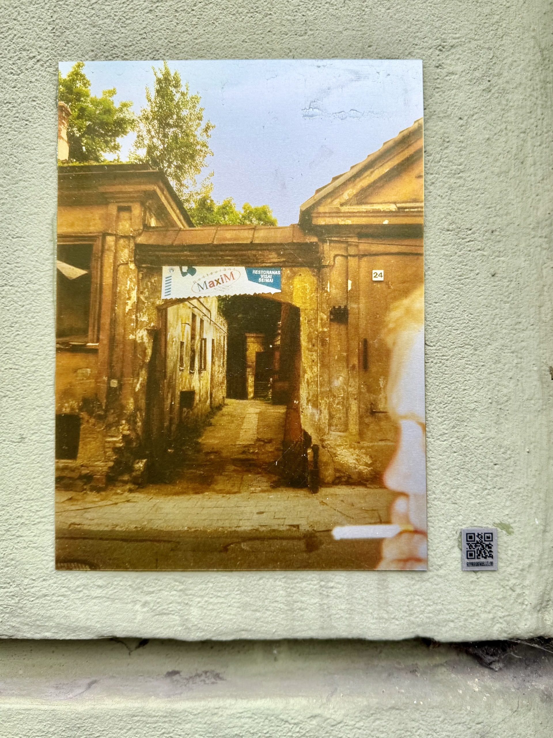 Archival photo of a courtyard entrance in Užupis An archival photograph mounted on a wall in Užupis showing the same courtyard entrance derelict, a restaurant banner strung across the arch.