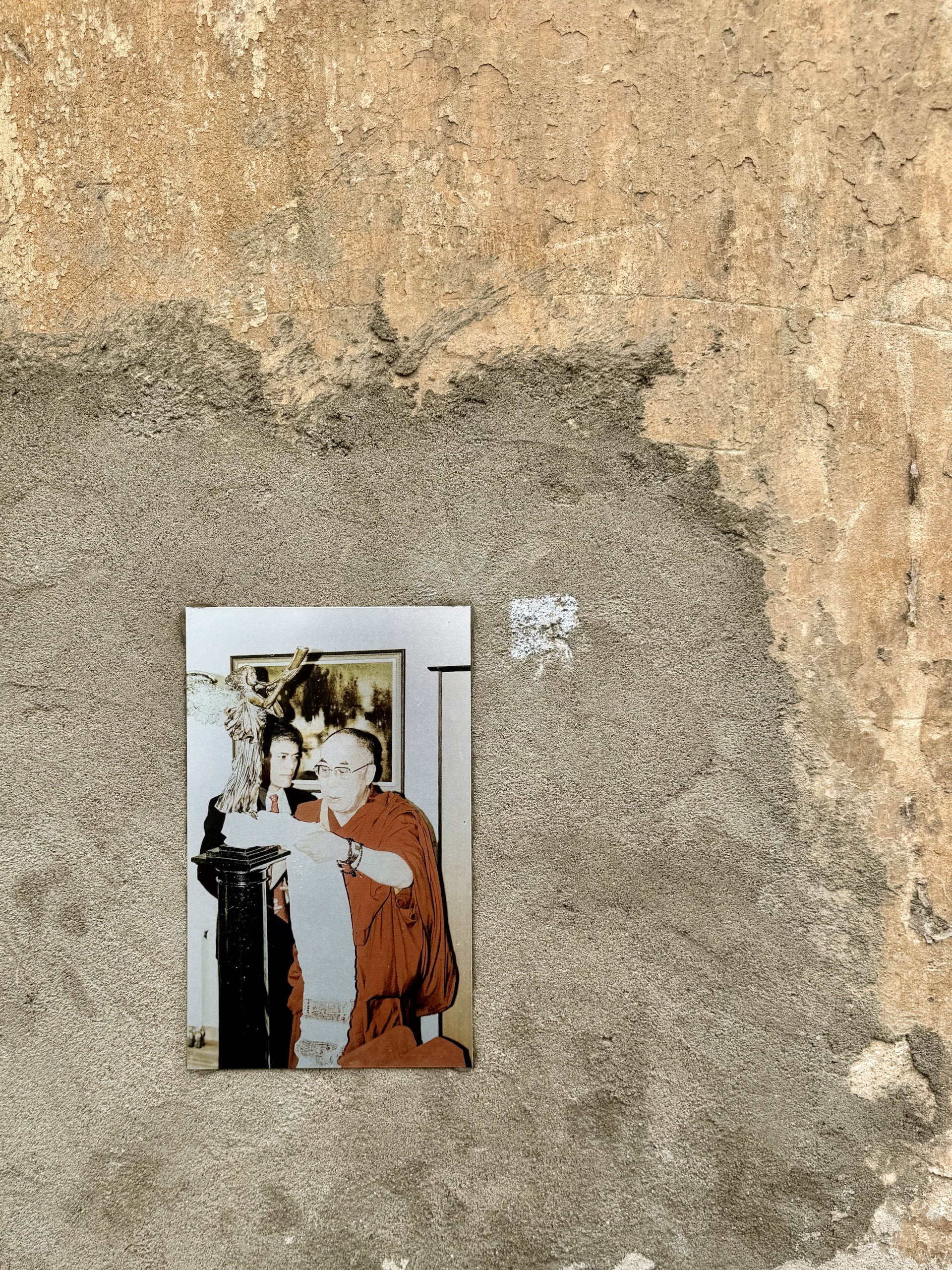 Archival photo of the Dalai Lama and the Angel of Užupis An archival photograph of the Dalai Lama beside a small model of the Angel of Užupis, mounted on a patch of bare cement render.