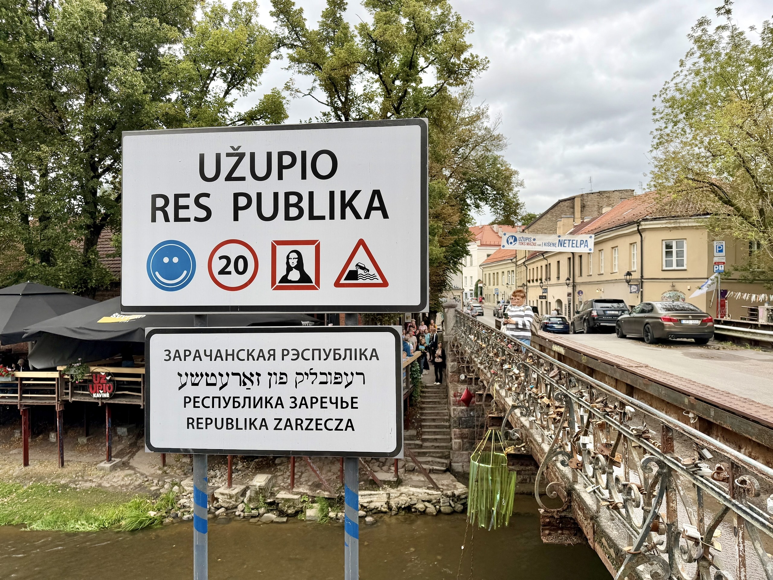 The Užupio Res Publika border sign on the bridge over the Vilnelė, with pictograms for a smile, a 20 km/h limit, the Mona Lisa and a car going into the river.