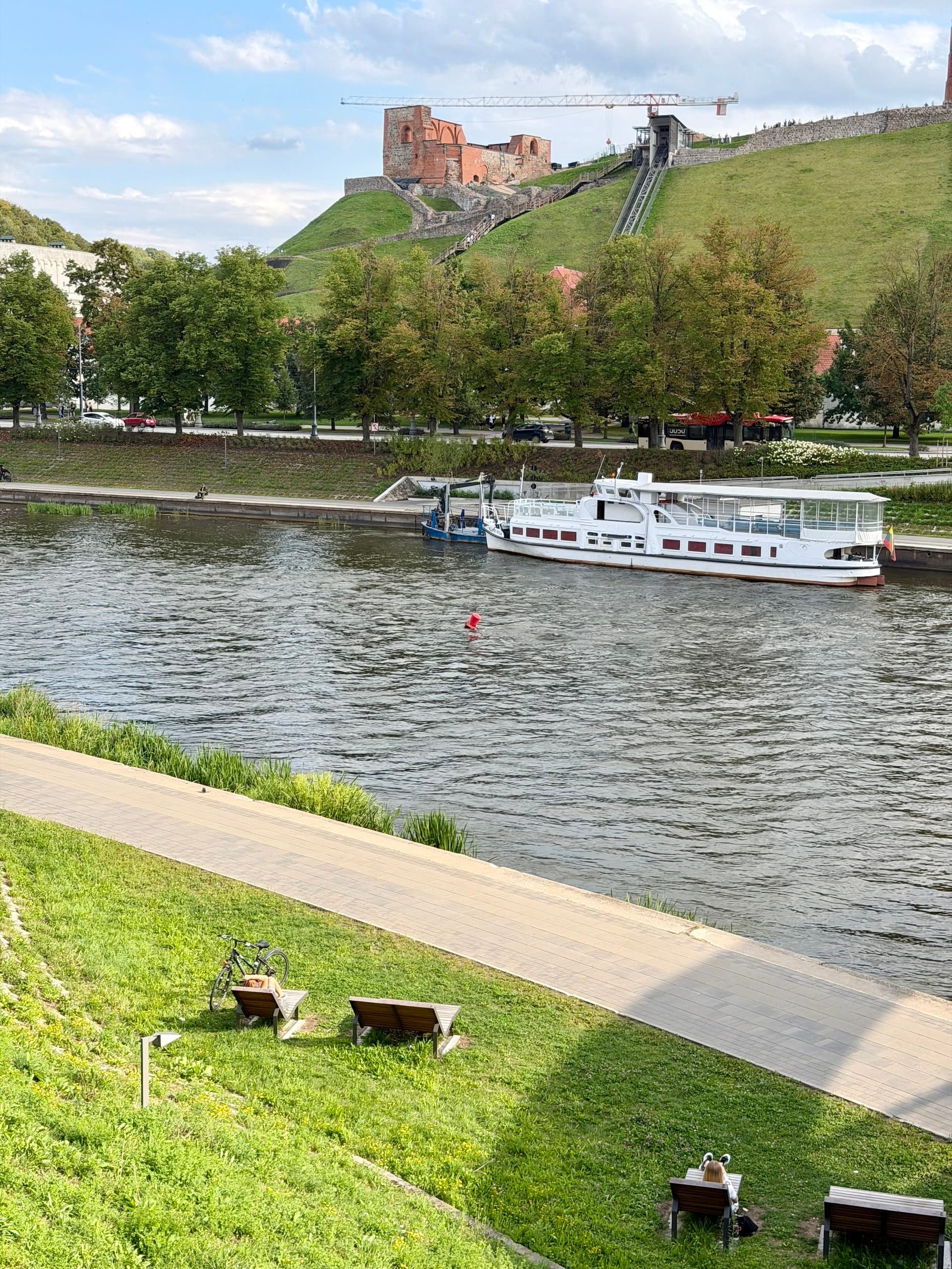 The bank of the Neris in Vilnius with Gediminas Hill behind Benches and lawns on the bank of the Neris in Vilnius, a river boat moored opposite and Gediminas Hill behind.