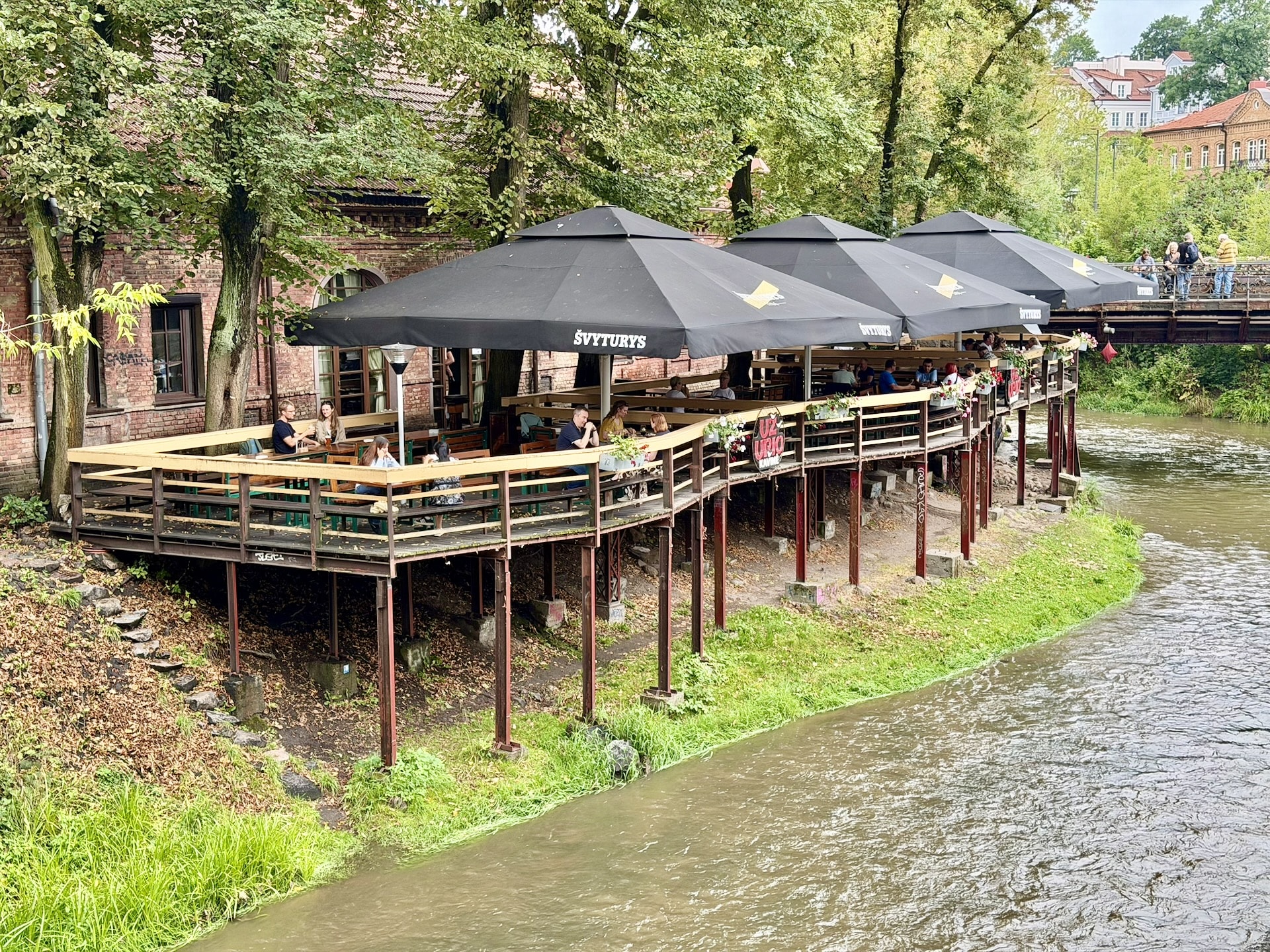 The Barlament bar on the Vilnelė in Užupis Riverside terrace bar on stilts above the Vilnelė in Užupis, Vilnius, black parasols over the tables and a red brick building behind.