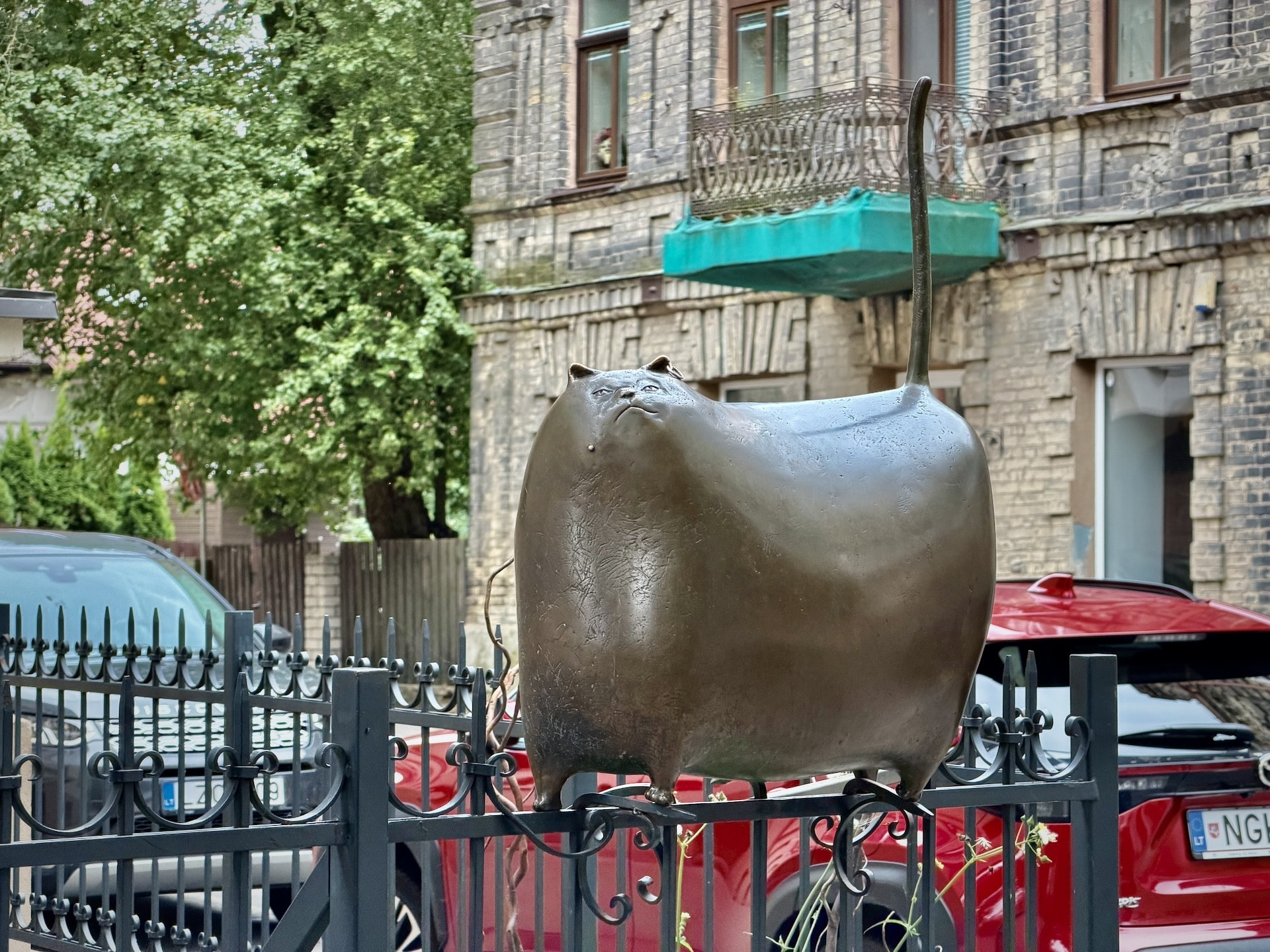 The Užupis Cat sculpture in Vilnius The bronze Užupis Cat sculpture on iron railings in Vilnius, an unrendered brick building and a rusting balcony behind it.