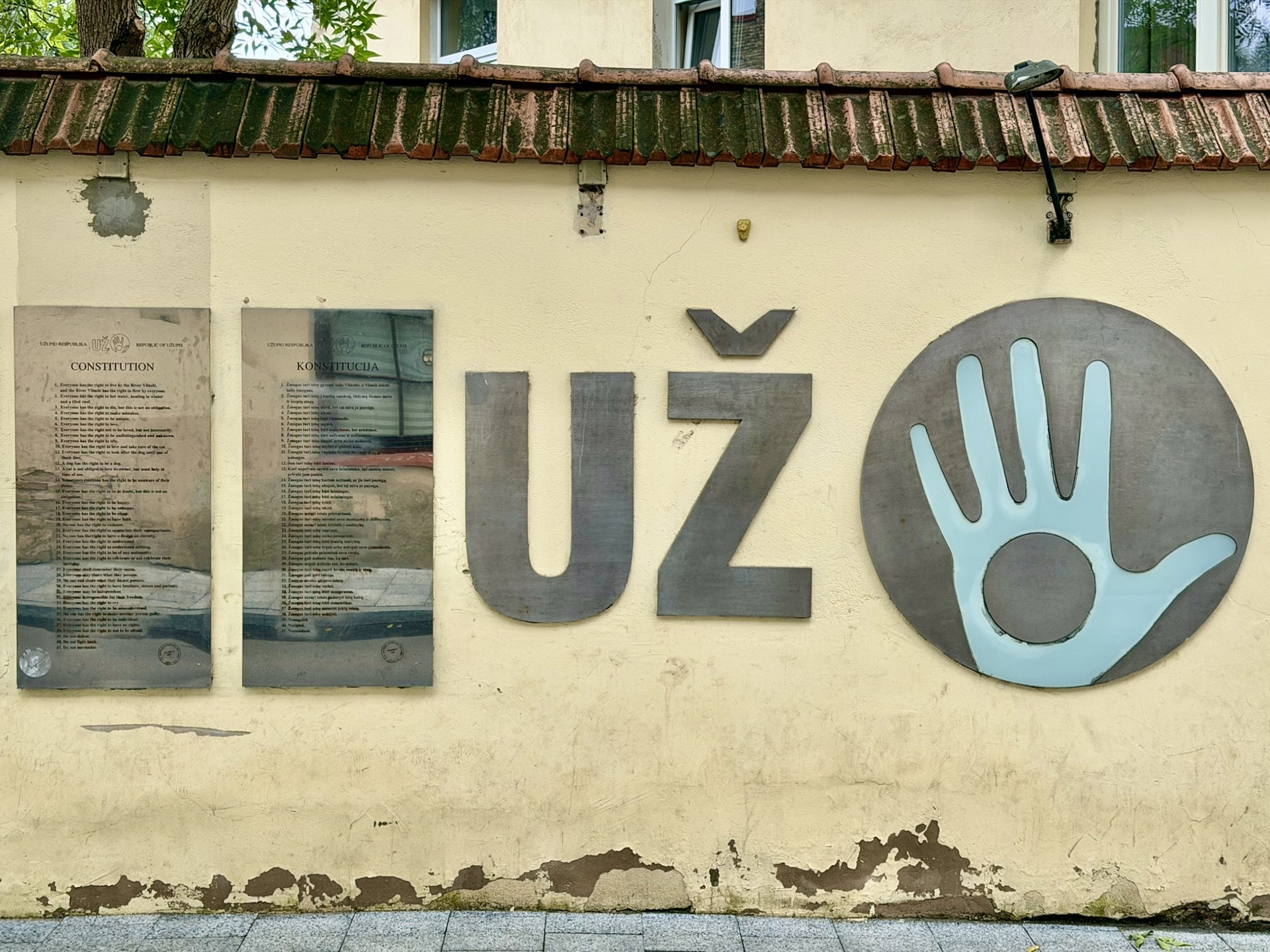 The Užupis constitution in Lithuanian and English Constitution plaques in English and Lithuanian on a yellow wall in Užupis, beside the UŽ lettering and the blue open-palm emblem of the republic.