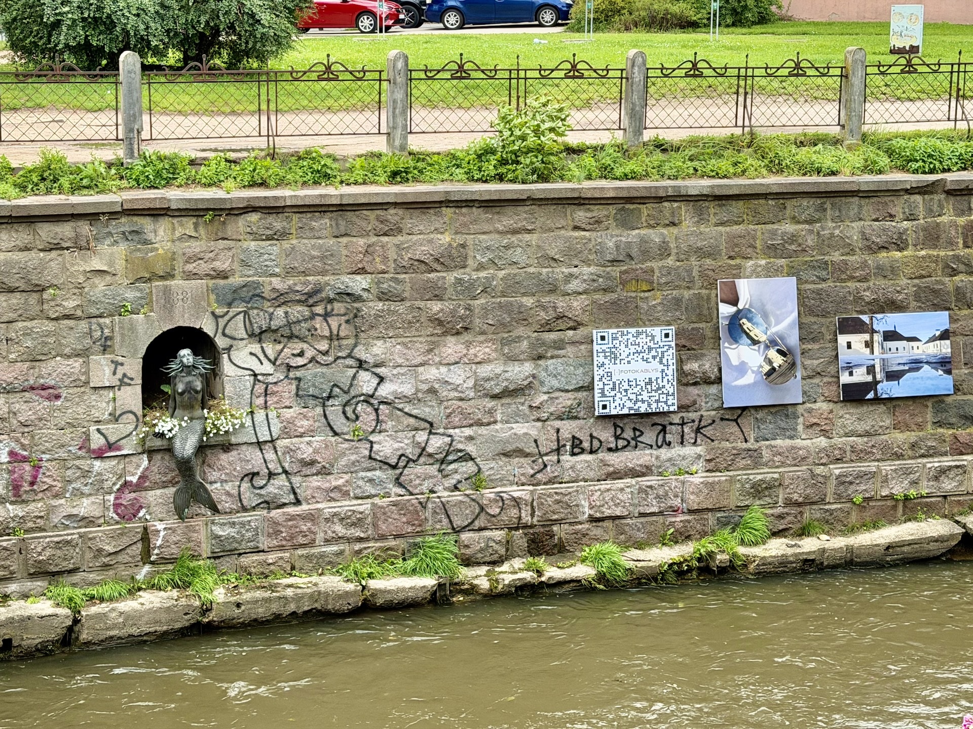 The Užupis Mermaid and the riverside exhibition The Užupis Mermaid in her niche in the Vilnelė embankment wall, graffiti across the stone and photographs from an open-air exhibition mounted alongside.