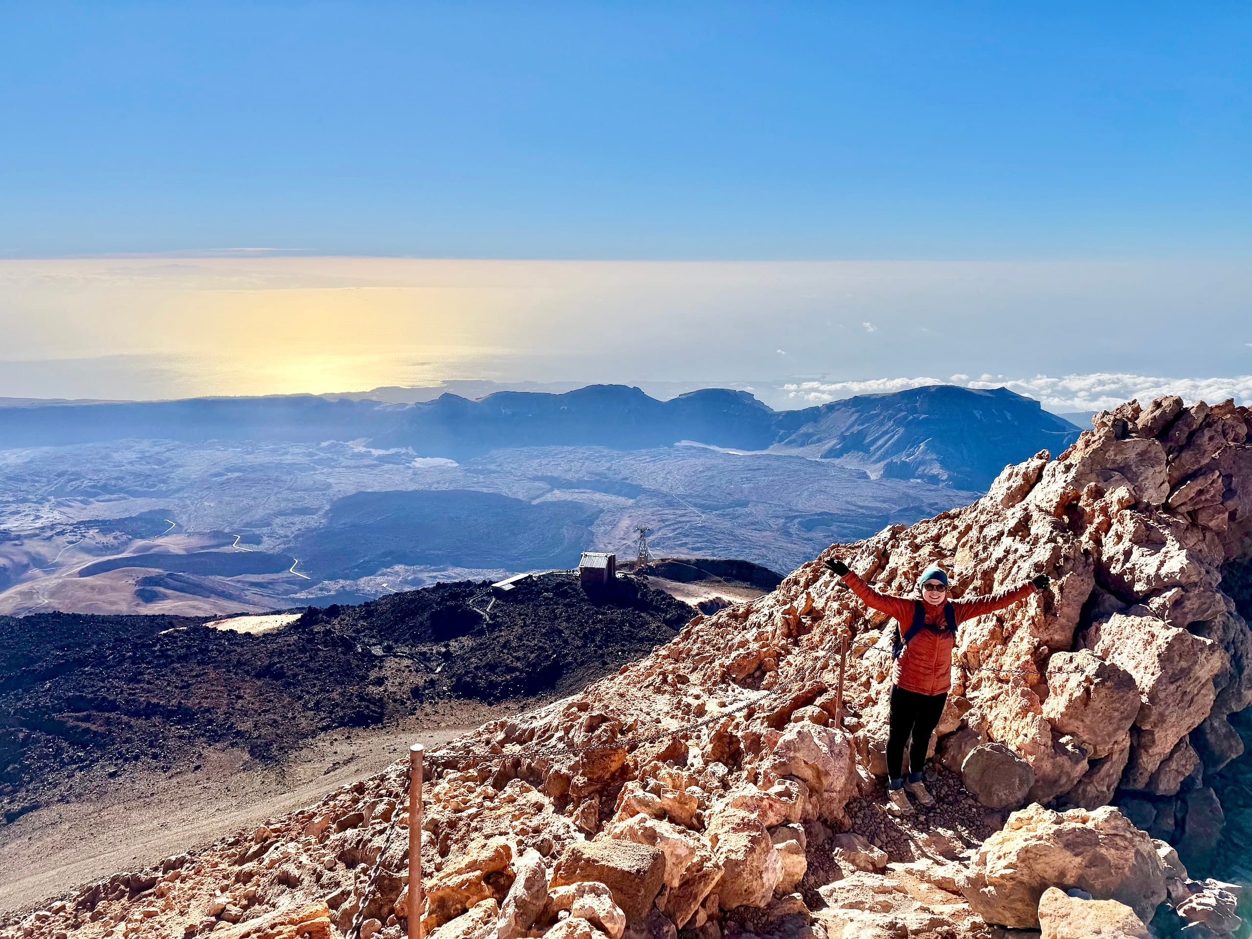 Andrea with her arms raised on the rocky slope near the summit of Mount Teide, the Las Cañadas caldera and the sea of clouds behind her