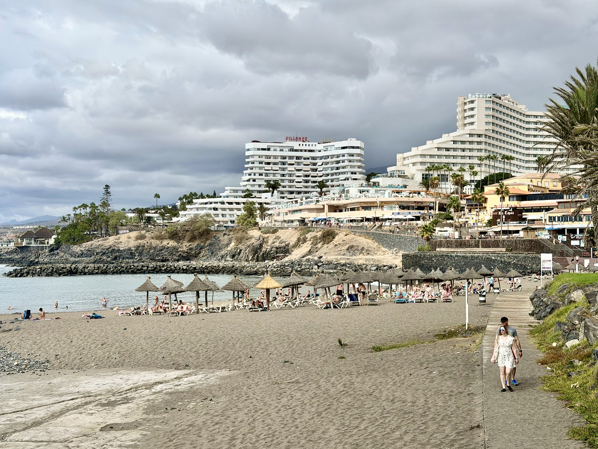 Grey sand beach with thatched parasols and sunbeds below large white hotel blocks in Costa Adeje, under heavy clouds, with a couple walking along the boardwalk