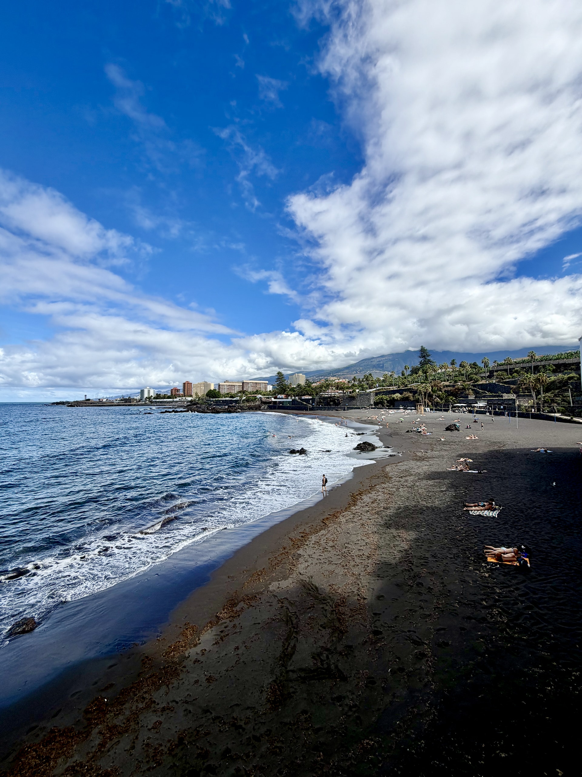Long black sand beach at Playa Punta Brava curving east towards Puerto de la Cruz, with waves, sunbathers and the mountains behind