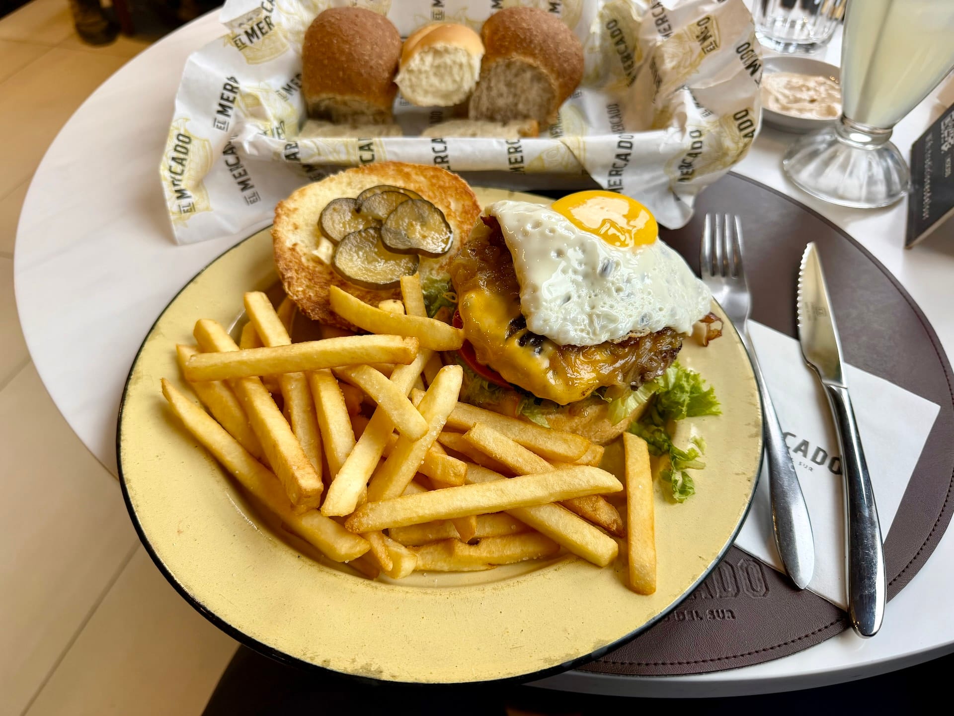 Burger topped with a fried egg and mushrooms, with fries and a bread basket, at El Mercado Ushuaia