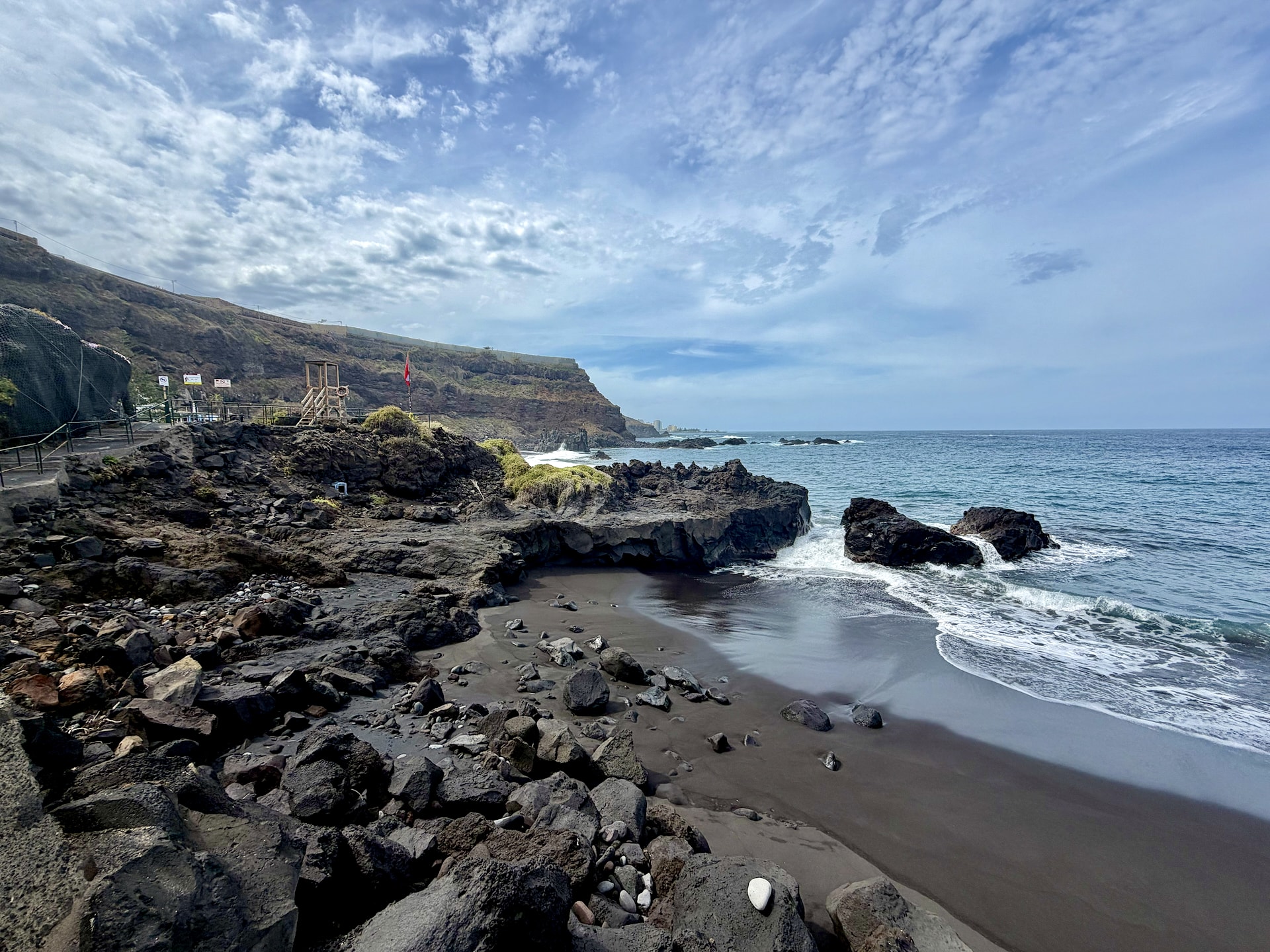 Closed section of the shore at El Bollullo, Tenerife Small black sand cove strewn with volcanic boulders beside El Bollullo, with a lifeguard tower and a red flag behind it