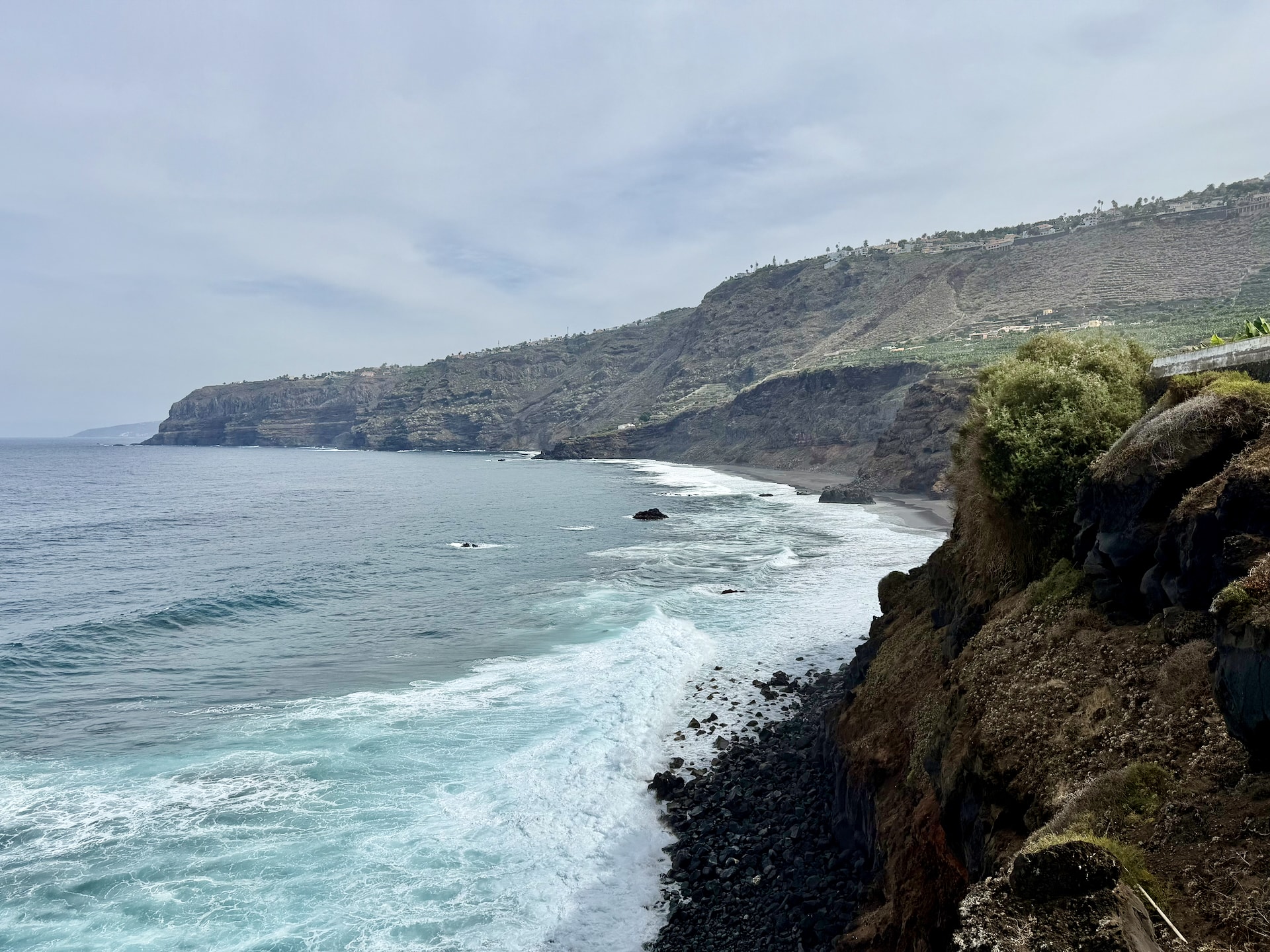 Coast toward Playa Los Patos from El Bollullo, Tenerife Surf breaking on a stony shore below steep cliffs east of El Bollullo, with Playa Los Patos in the distance and houses along the clifftop