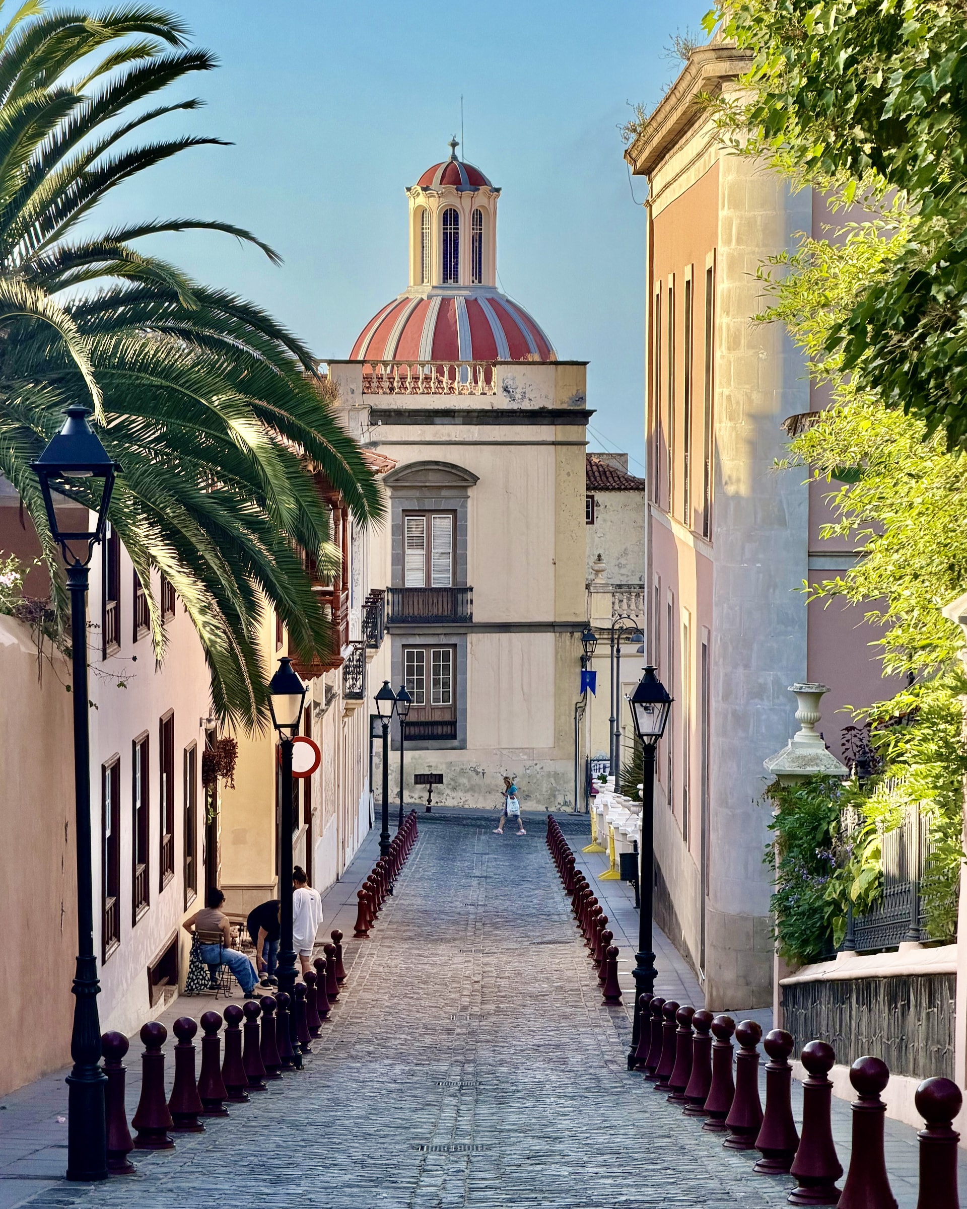 Cobbled street in La Orotava's old town lined with maroon bollards, iron lamps and a palm tree, leading to a red and white striped church dome