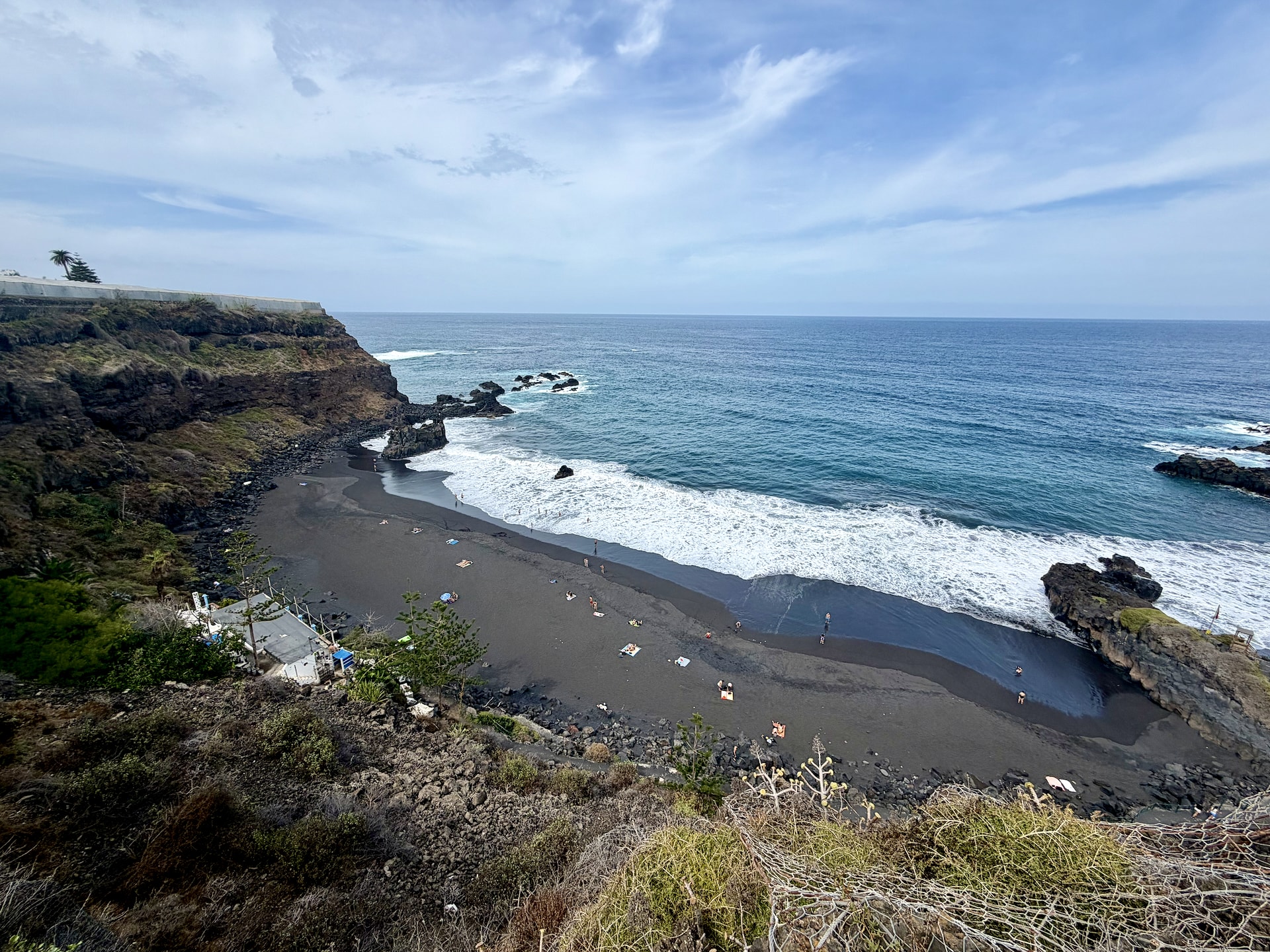 El Bollullo black sand beach and its café, seen from the cliff El Bollullo from the cliff path: black sand with scattered sunbathers, white surf, offshore rocks and the beach café's buildings at the foot of the cliff