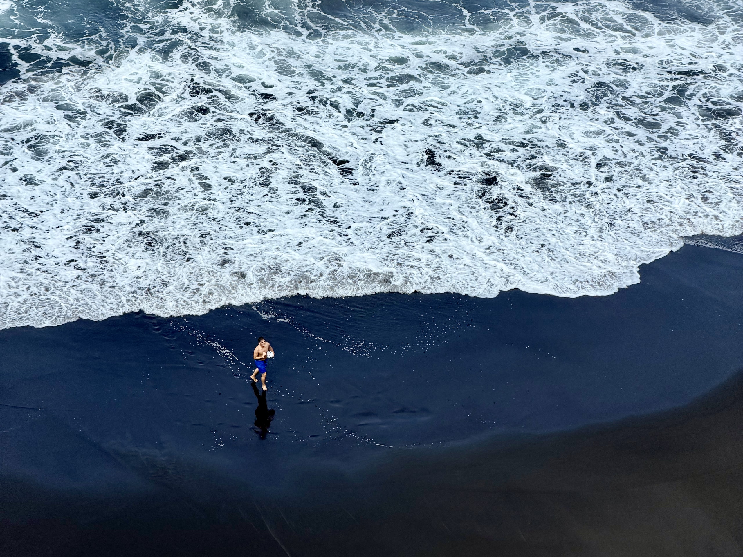 Aerial view of El Bollullo beach in Tenerife: a man in blue swim shorts carrying a ball across the black sand as white surf rolls in