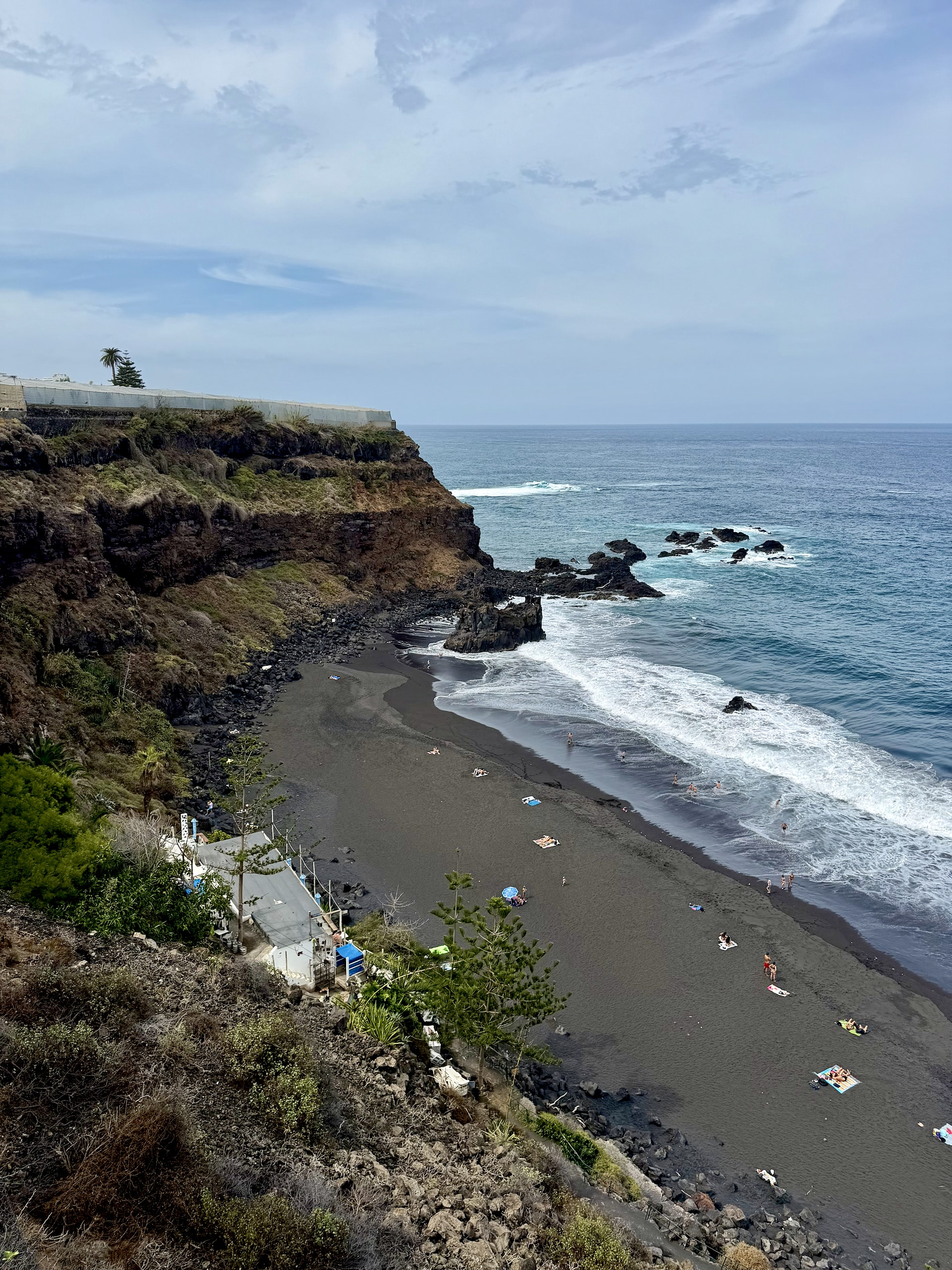 El Bollullo black sand beach seen from the cliff El Bollullo from the cliff path above: a strip of black sand at the foot of green cliffs, with sunbathers, the beach bar and white surf