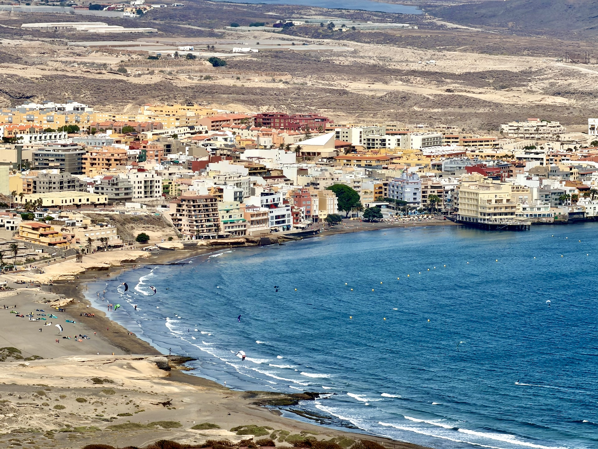 El Médano bay and kitesurfers seen from Montaña Roja El Médano seen from Montaña Roja: a curved bay with kitesurfers' kites over the water, the beach and the town's apartment buildings against dry hills