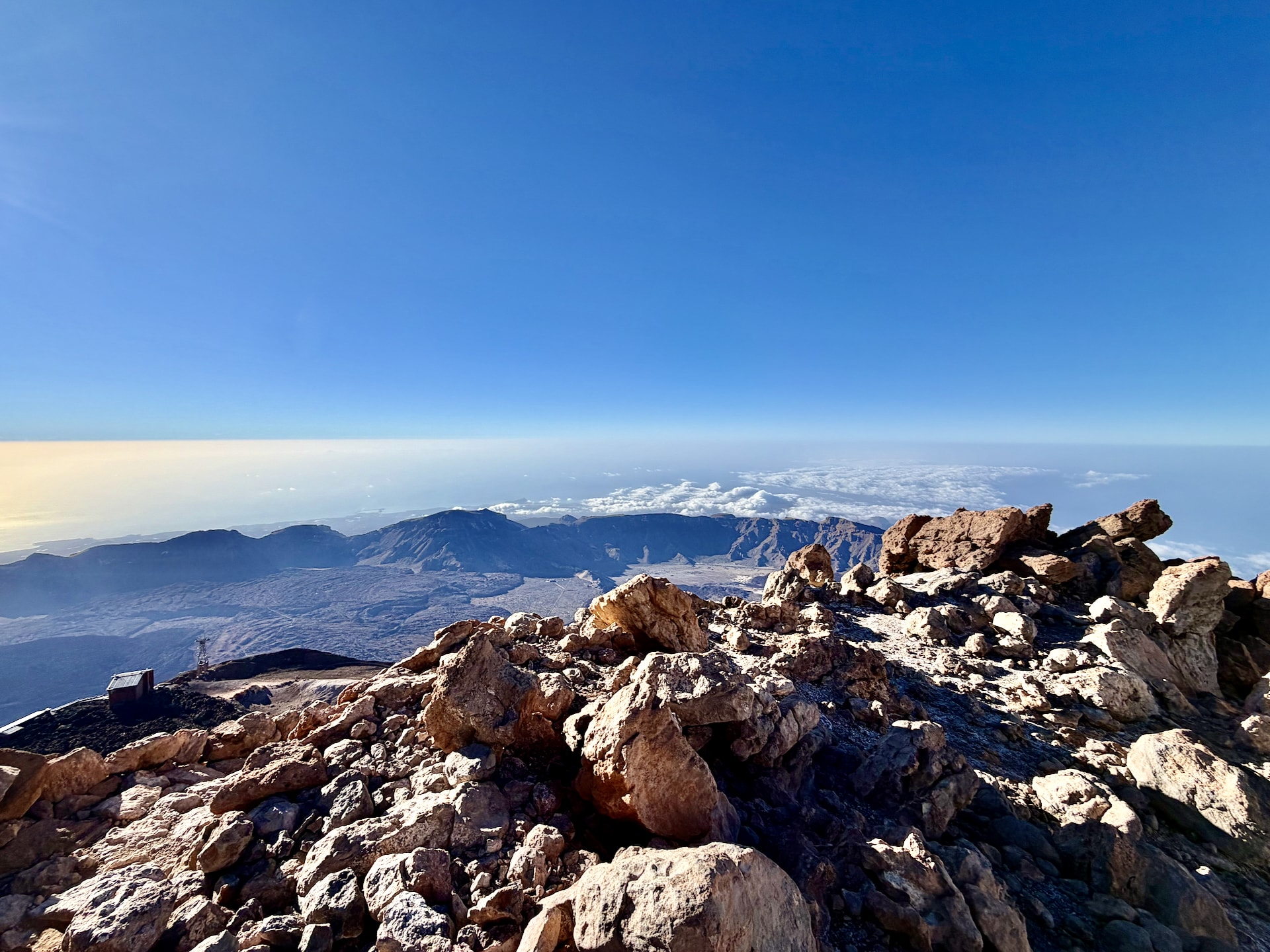 Jagged volcanic rocks at the top of Mount Teide in the foreground, with the caldera, distant mountains and a layer of clouds beyond