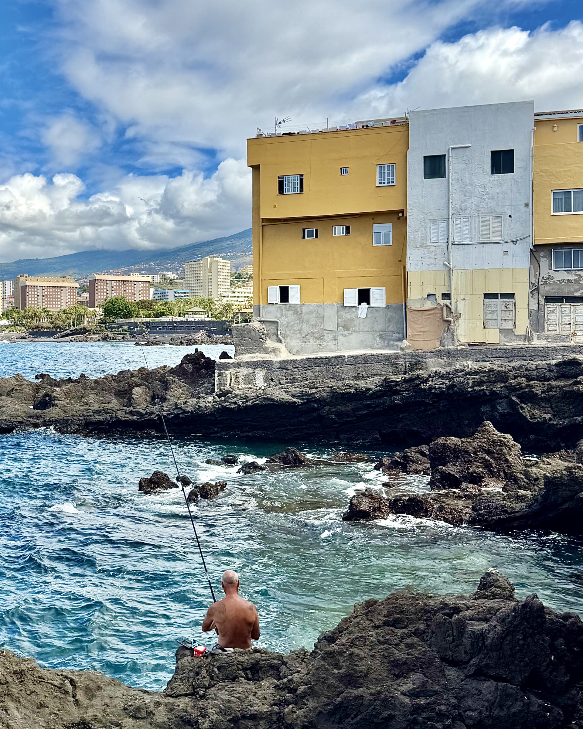 Fisherman casting from black lava rocks in front of yellow and white houses on the Punta Brava seafront