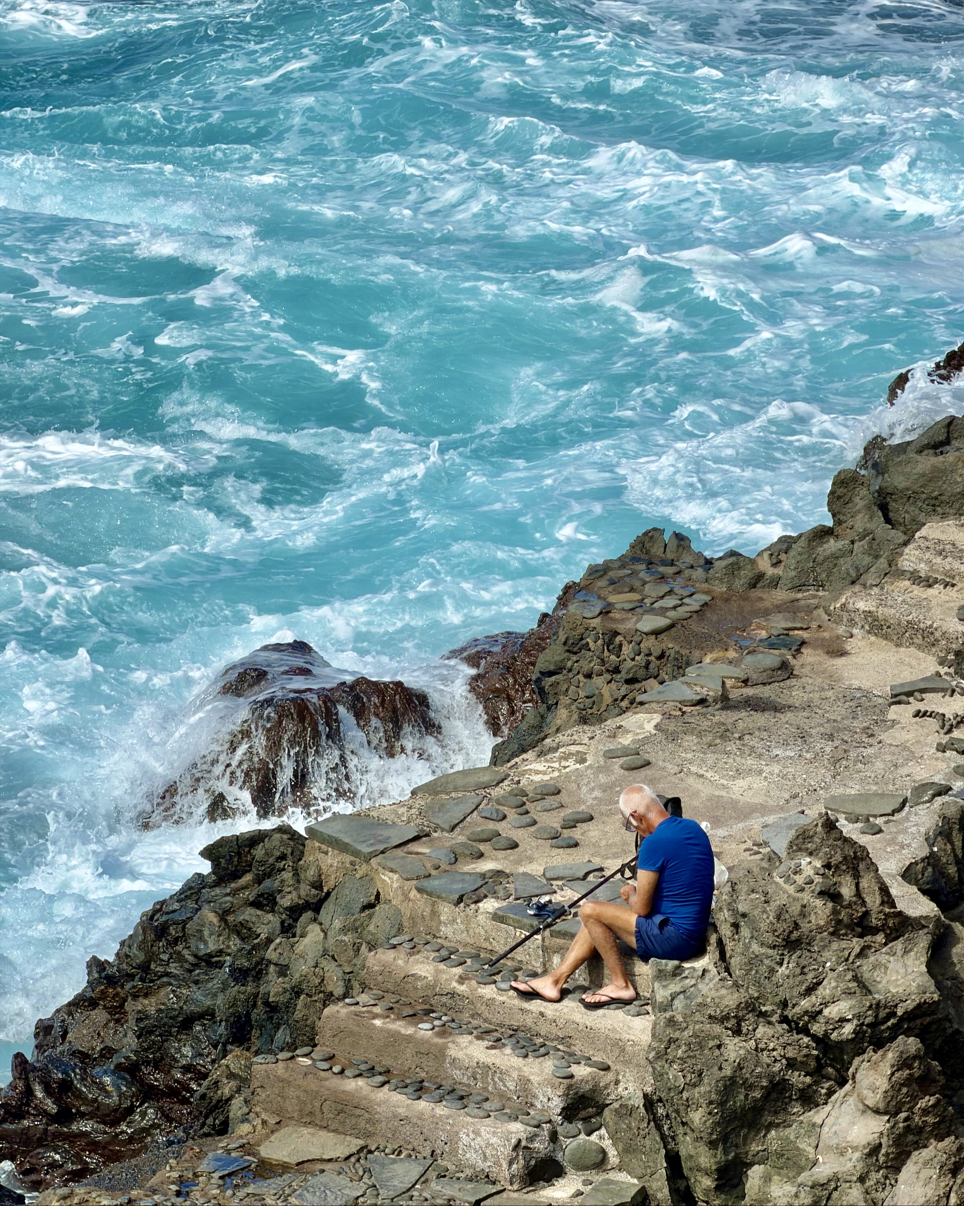 Fisherman in a blue shirt sitting on stone steps above crashing turquoise waves at Punta Brava
