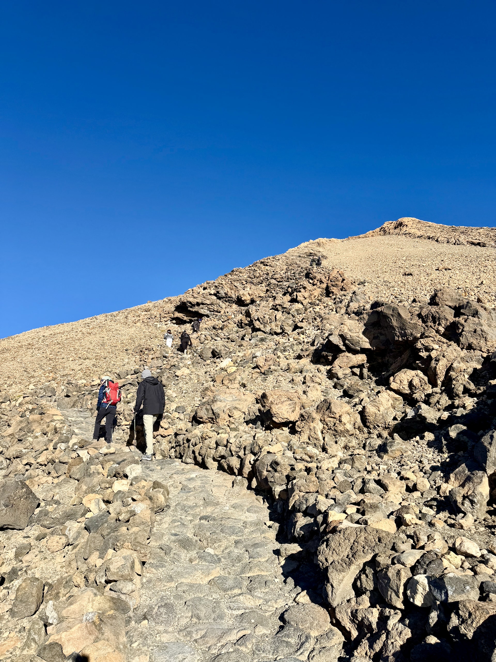 Two hikers walking up the stone-paved summit trail on Mount Teide between loose volcanic boulders under a deep blue sky