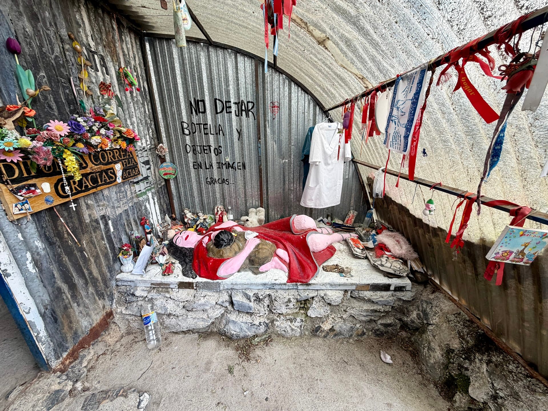 Inside a Difunta Correa roadside shrine near Ushuaia, red ribbons, flowers and offerings under a corrugated metal roof