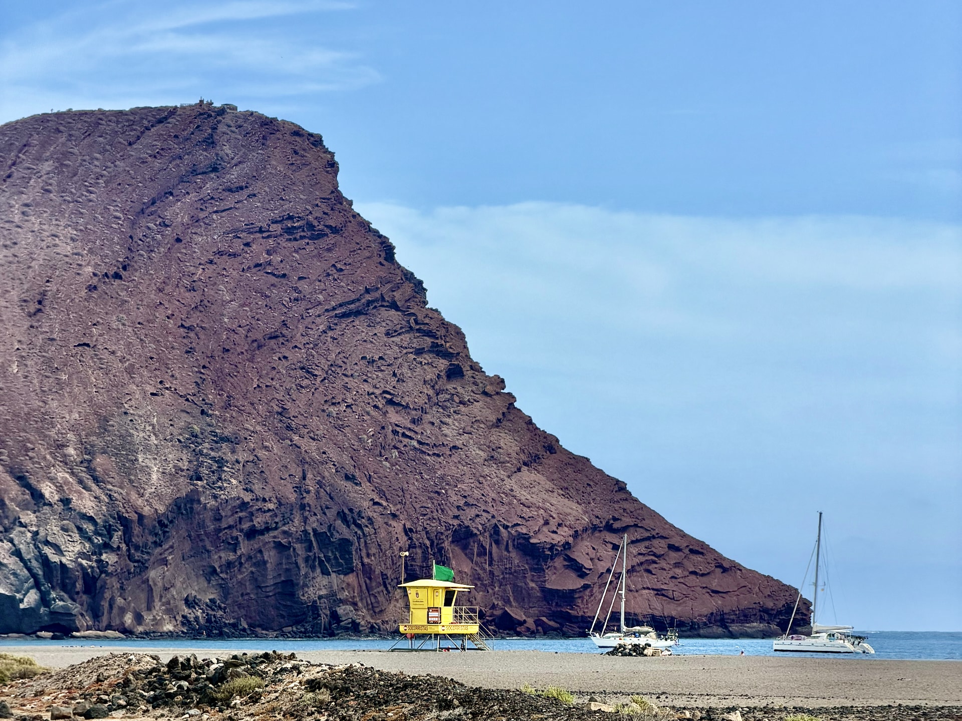 La Tejita beach and Montaña Roja volcano, south Tenerife Red-brown slope of Montaña Roja rising behind La Tejita beach, with a yellow lifeguard tower and two sailboats anchored offshore