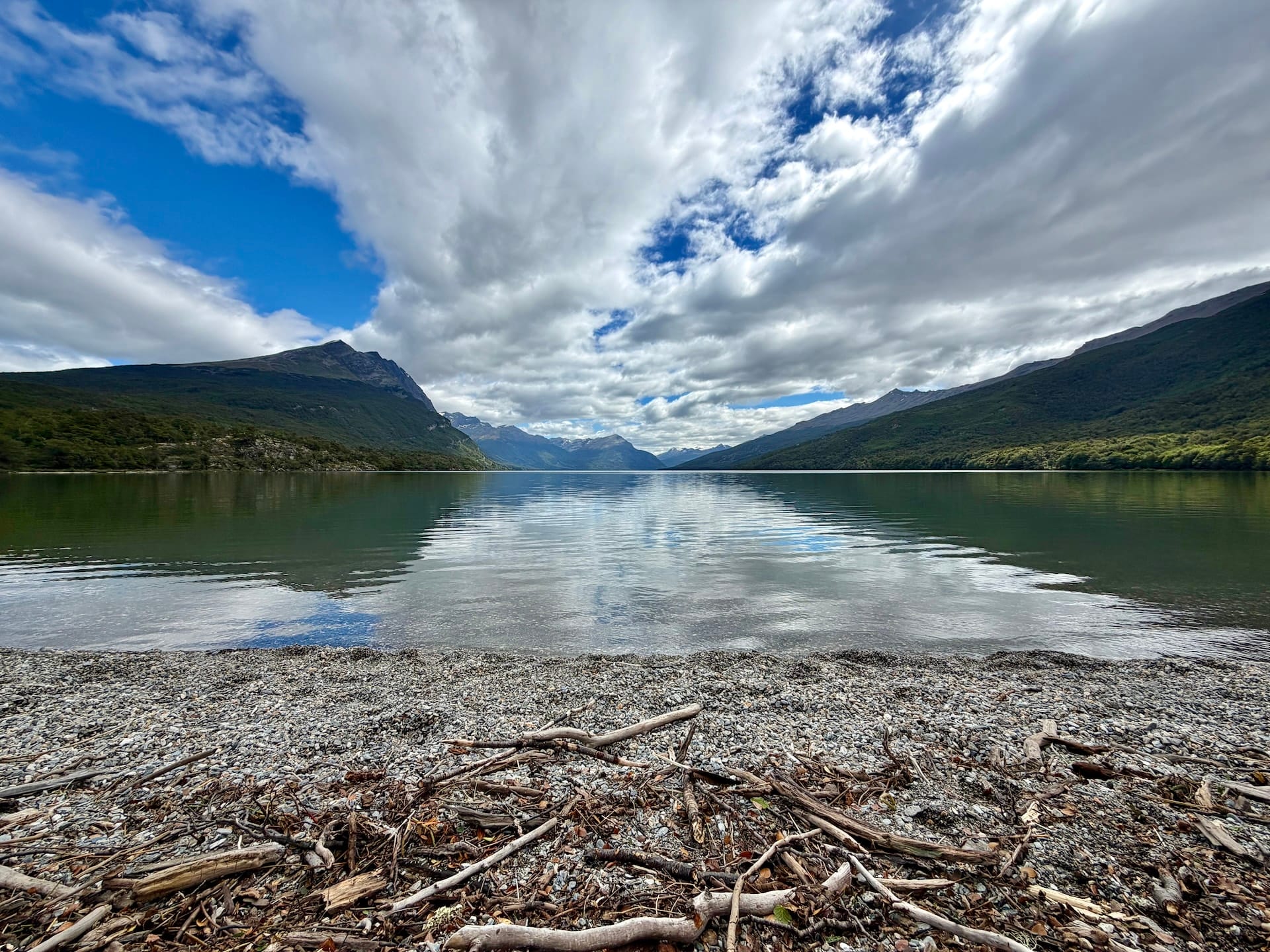 Calm Lago Roca reflecting clouds and forested mountains, driftwood on the pebble shore