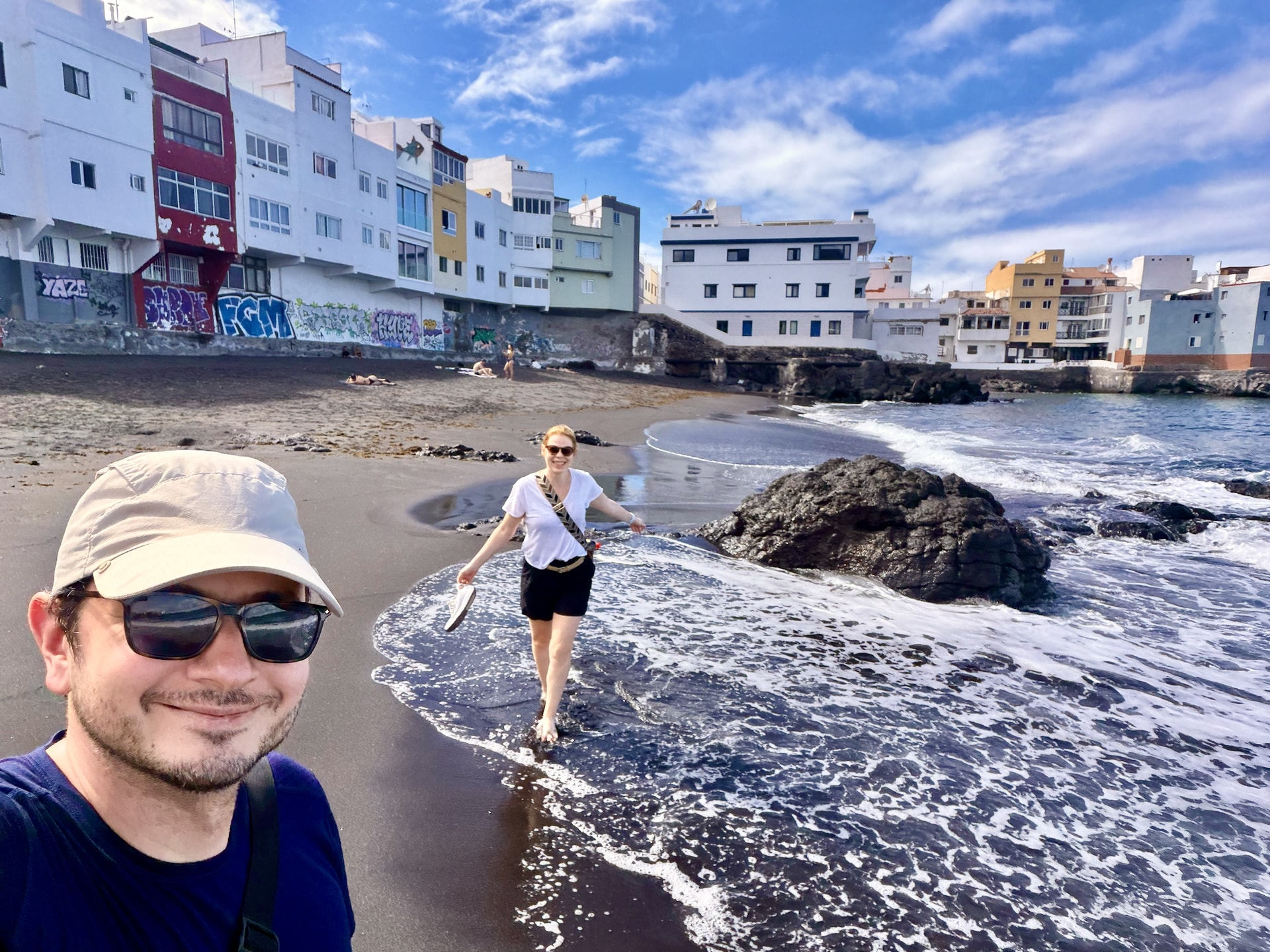 Laszlo taking a selfie on the black sand of Playa Punta Brava as Andrea wades barefoot through the surf, with colorful houses and graffiti behind