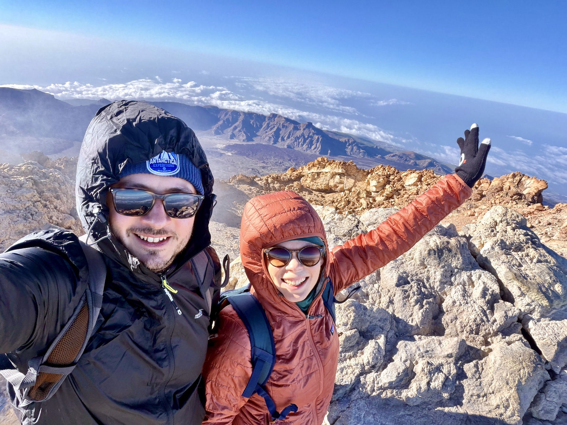 Laszlo and Andrea taking a selfie in warm jackets and gloves on the summit of Mount Teide, with the caldera and clouds far below
