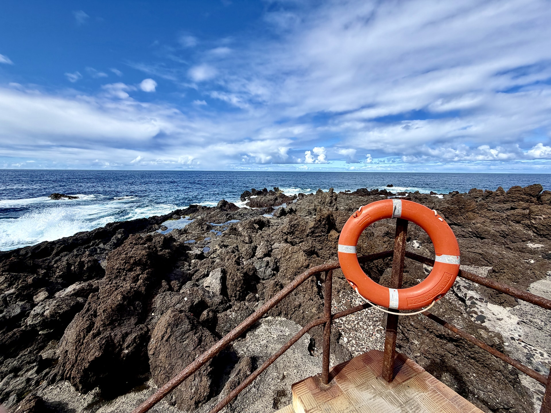 Orange life ring on a rusty railing above jagged black lava rocks and rock pools at Punta Brava