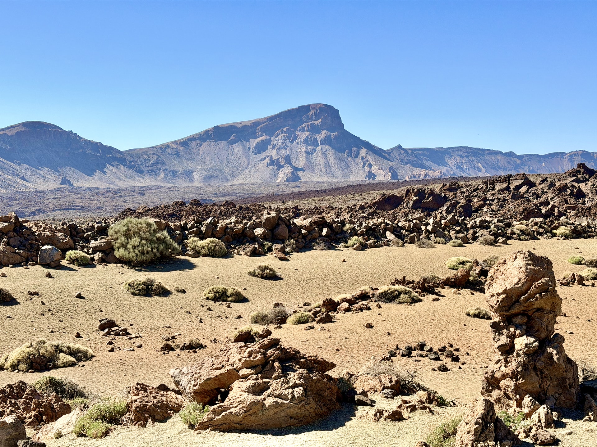Pale volcanic sand dotted with scrub and eroded rock pillars at Mirador de Minas de San José, with mountains in the background