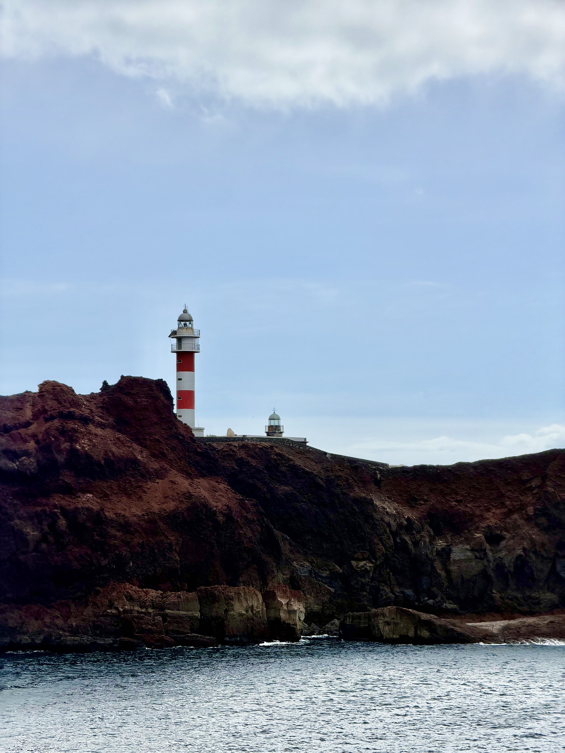 Red-and-white Punta de Teno lighthouse with the small old lighthouse beside it on a red volcanic cliff above the sea