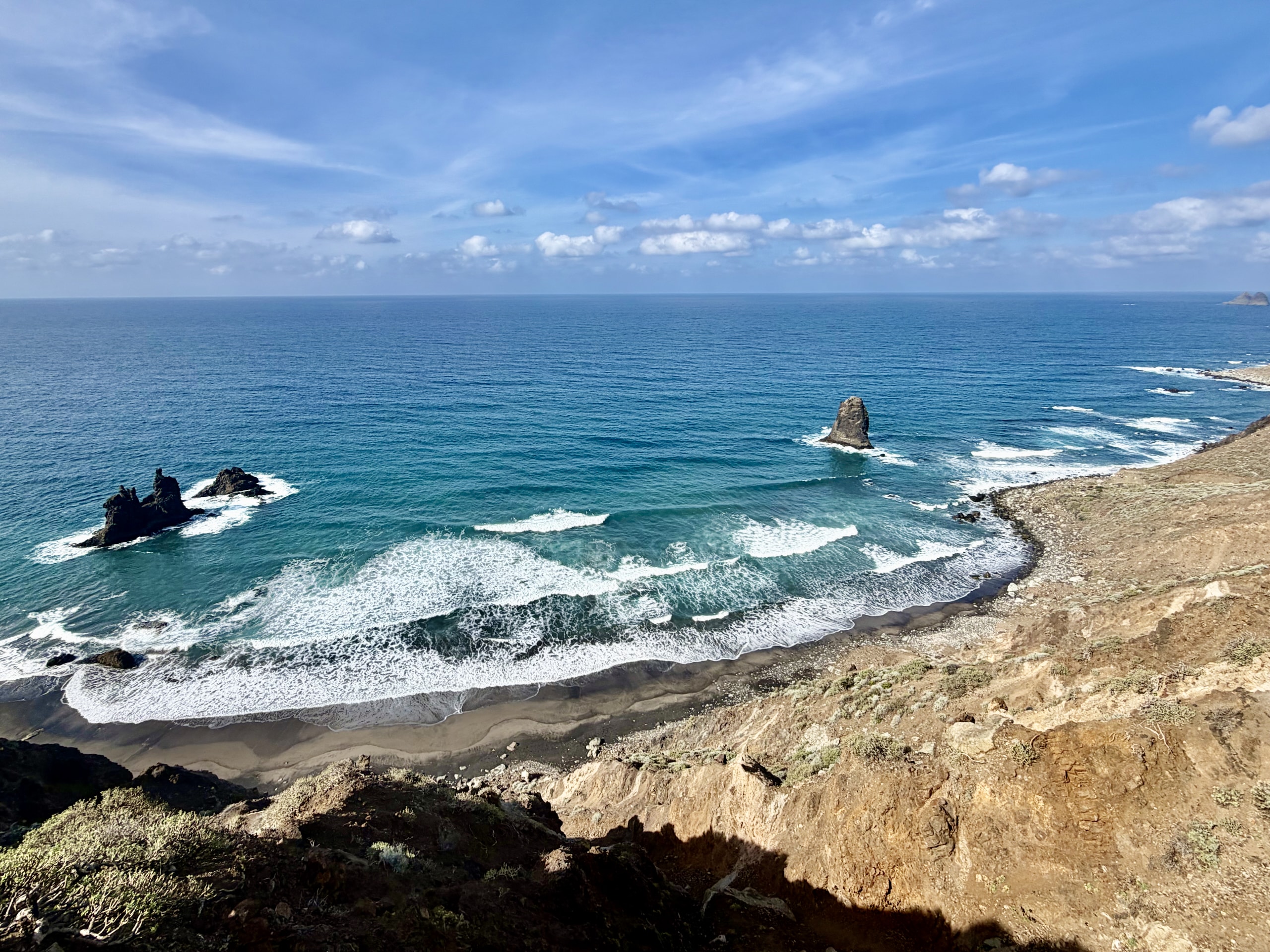 Playa de Benijo seen from the coastal hiking trail: a dark sand beach, white surf and sea stacks off the Anaga coast under a blue sky